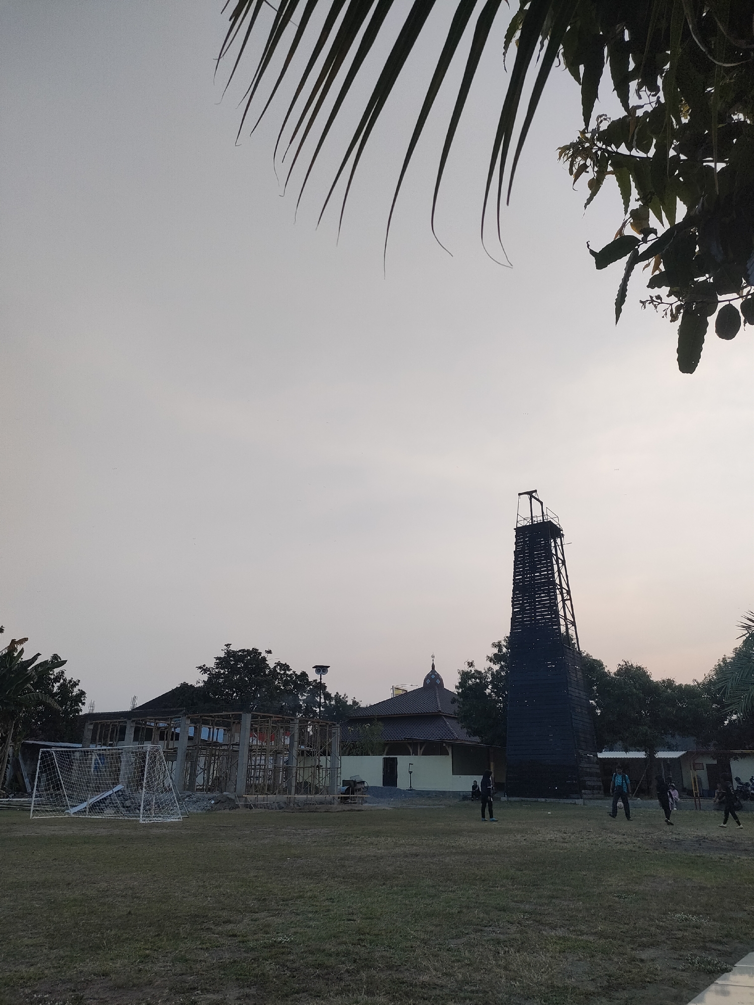 The image depicts an outdoor scene at dusk in Banguntapan, Indonesia. In the foreground, a grassy field shows a soccer goal on the left. In the middle ground, a building under construction is visible, with scaffolding and structural elements. To the right of the construction site, a tall, dark wooden tower stands prominently. Behind the tower, a building with a dome, possibly a mosque, is partially obscured by trees. Several individuals are scattered across the field, some appearing to be playing soccer. The sky is overcast, and the overall lighting suggests the end of the day. Trees and foliage are present on the sides and in the background, framing the scene.