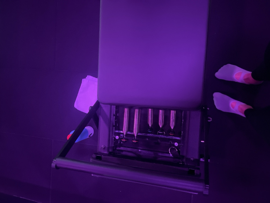 The image shows a pilates chair bathed in purple light. The chair has a padded seat and four visible springs beneath it. The springs appear silver. A white towel is draped over the side of the chair, and a small cylindrical object rests beside it. To the right of the chair are two feet wearing white socks with pink accents, presumably belonging to someone using the equipment. The floor and surrounding walls are a dark, contrasting shade of purple.