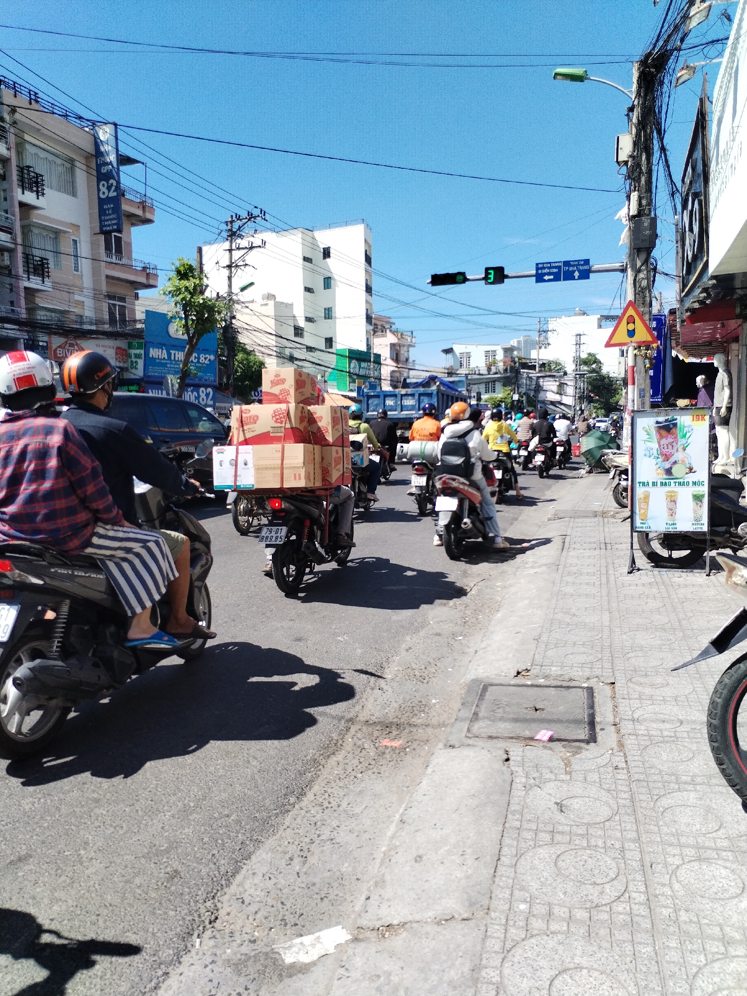 This image captures a busy street scene in Nha Trang, Vietnam, specifically in the Bac Nha Trang Ward. A two-lane road is filled with a convoy of motorcycles, many carrying passengers and goods. In the foreground, two individuals are riding a motorcycle, with the rider wearing a plaid shirt and striped pants and the passenger wearing a helmet. Behind them, another motorcycle is laden with stacked boxes, likely merchandise. The road is lined with buildings, including shops and multi-story structures, with overhead power lines and traffic signals visible. The atmosphere is dynamic, suggesting active commerce and daily life in the city.