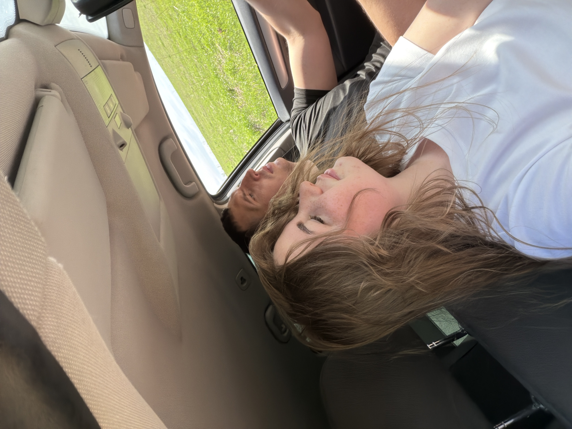 A woman with fair skin and freckles, wearing a white t-shirt, and a man with tan skin and wearing a black t-shirt, are lying in a car with their heads near the car's sunroof. The woman's light brown hair is spread out over the car seat. The car is moving, as can be seen by the green grass visible through the sunroof. The location, Ural'skoe, Russia, cannot be confirmed from this image alone. The image shows the interior of a car with the sunroof open.