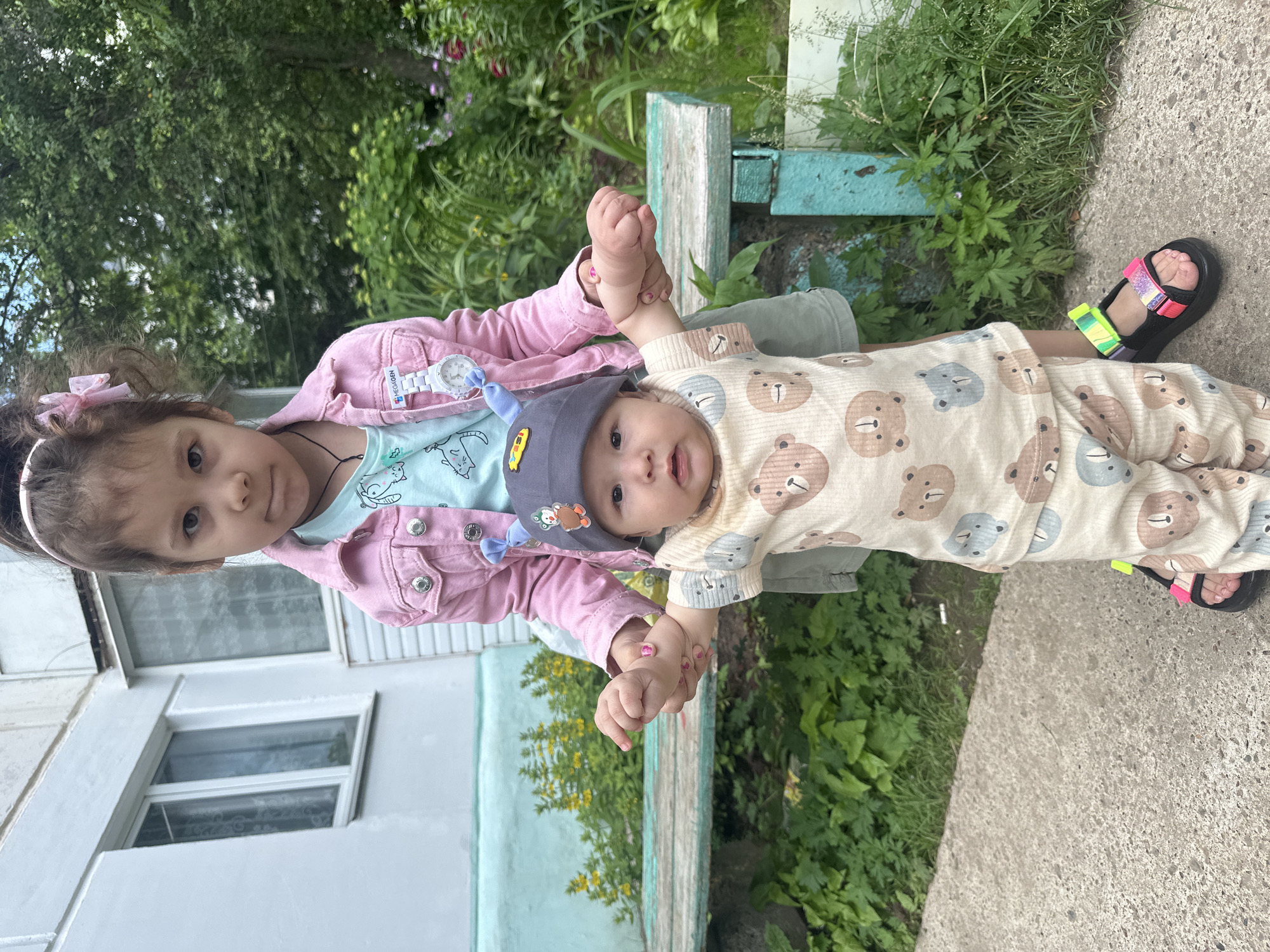 Two young children are standing on a concrete pathway in front of a light-colored building and a bench. The taller child, a girl with dark hair tied up with a pink bow, wears a pink denim jacket over a light blue shirt. She is holding the hand of the younger child, a baby, who is wearing a gray hat with blue embellishments and a light-colored outfit with a pattern of bears. The baby's arms are extended forward. The background shows greenery and part of a building with windows. The overall scene appears to be outdoors in a residential area.