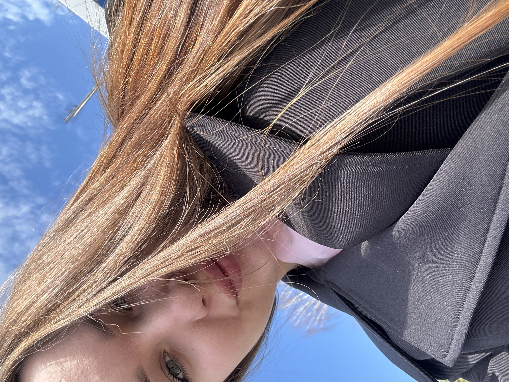 The scene is situated outdoors in the city of Noviy, Russia. The image is a close-up, low-angle, self-portrait capturing a young female against a bright blue sky with scattered white clouds. The subject's long, light brown hair dominates the right side of the frame, flowing across her face and the dark jacket she is wearing. Her left eye, visible on the upper left, is green. Her nose and lips are partially visible, with a neutral expression. She wears a dark-colored jacket or trench coat with a lapel, over a light pink garment. In the distant, out-of-focus background against the sky, a slender light-colored pole or mast and a blurred white structure are discernible. Sunlight illuminates the subject's hair and clothing.