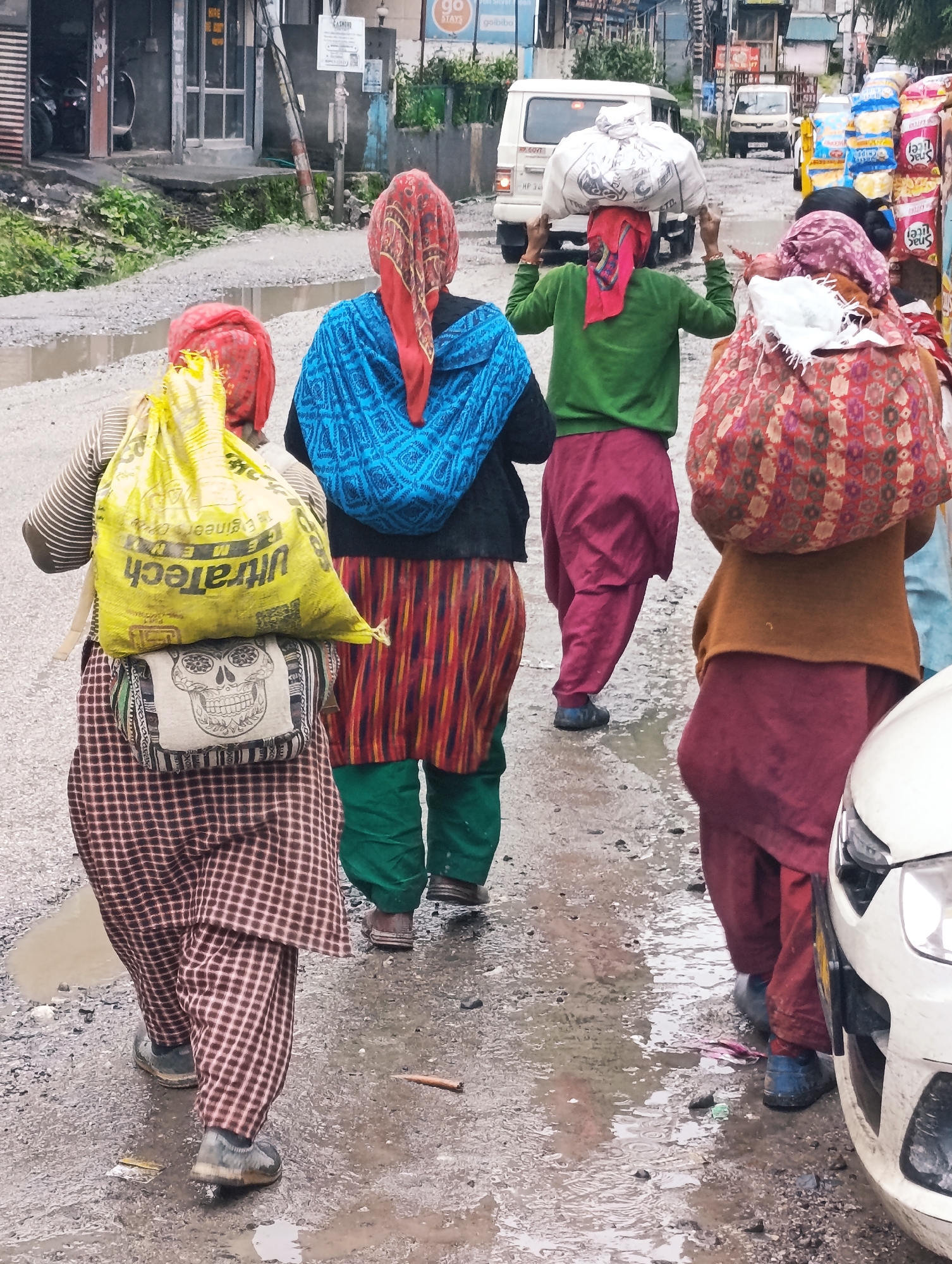 In Chichoga, India, four women are walking on a wet, gravelly road. The woman in the foreground, on the left, wears a red and yellow checkered salwar kameez and a yellow bag with "UltraTech" printed on it on her back. Below the yellow bag is a smaller bag with a skull print. The second woman from the left wears a blue and white patterned dupatta and a red and yellow striped skirt. The third woman from the left wears a green top and red salwar. She carries a white sack on her head. The woman on the far right wears a maroon salwar kameez and a patterned red and brown bag on her head. A white car is parked on the right side of the road. The background shows buildings and vehicles, including two white vans.