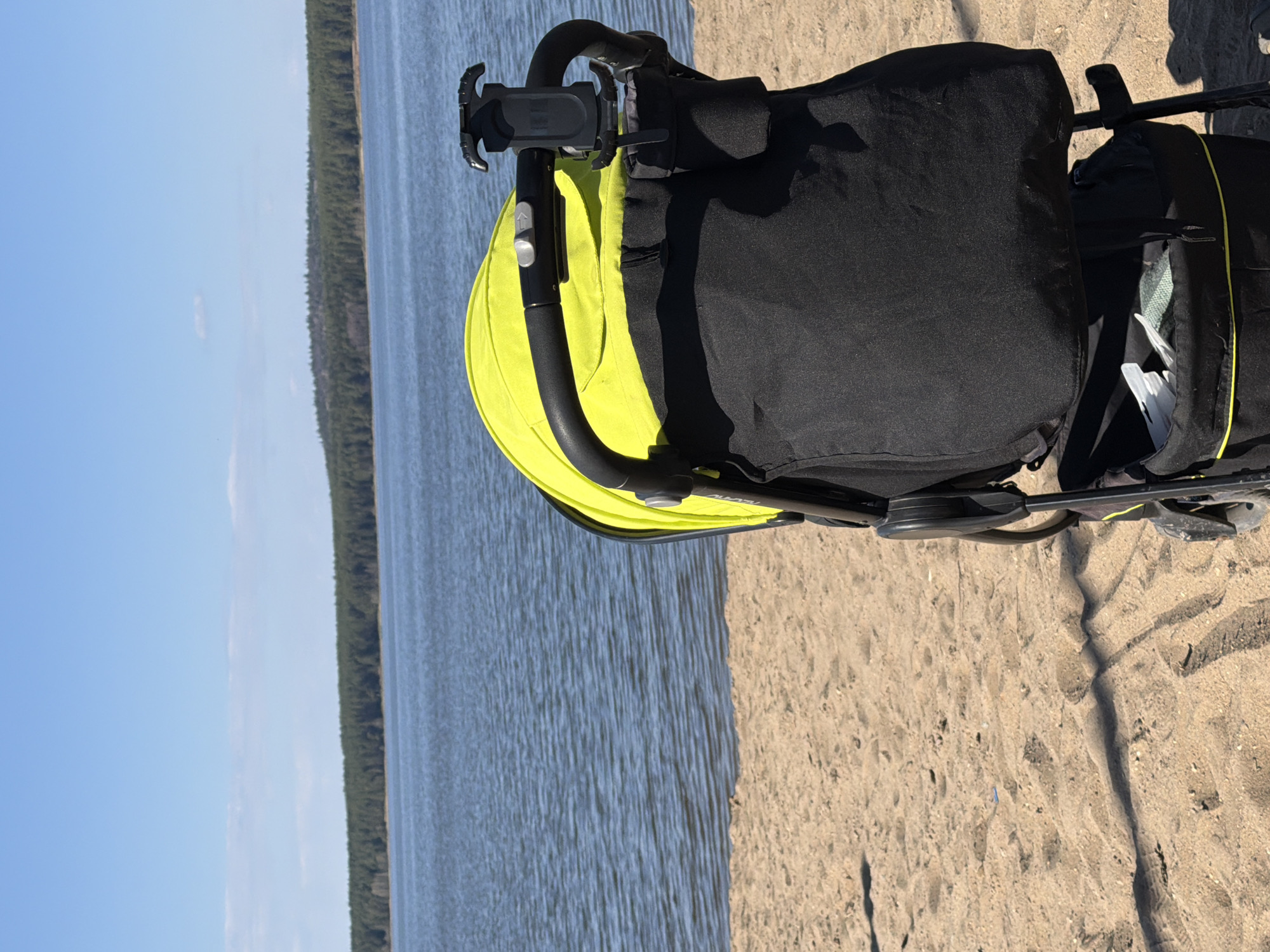 The image shows a stroller with a yellow sunshade and a black seat, parked on a sandy surface. Behind the stroller is a body of water, possibly a lake or river, with a forested shoreline on the opposite bank. The sky is blue with some clouds. The location is possibly near the city of Votkinsk, Russia.