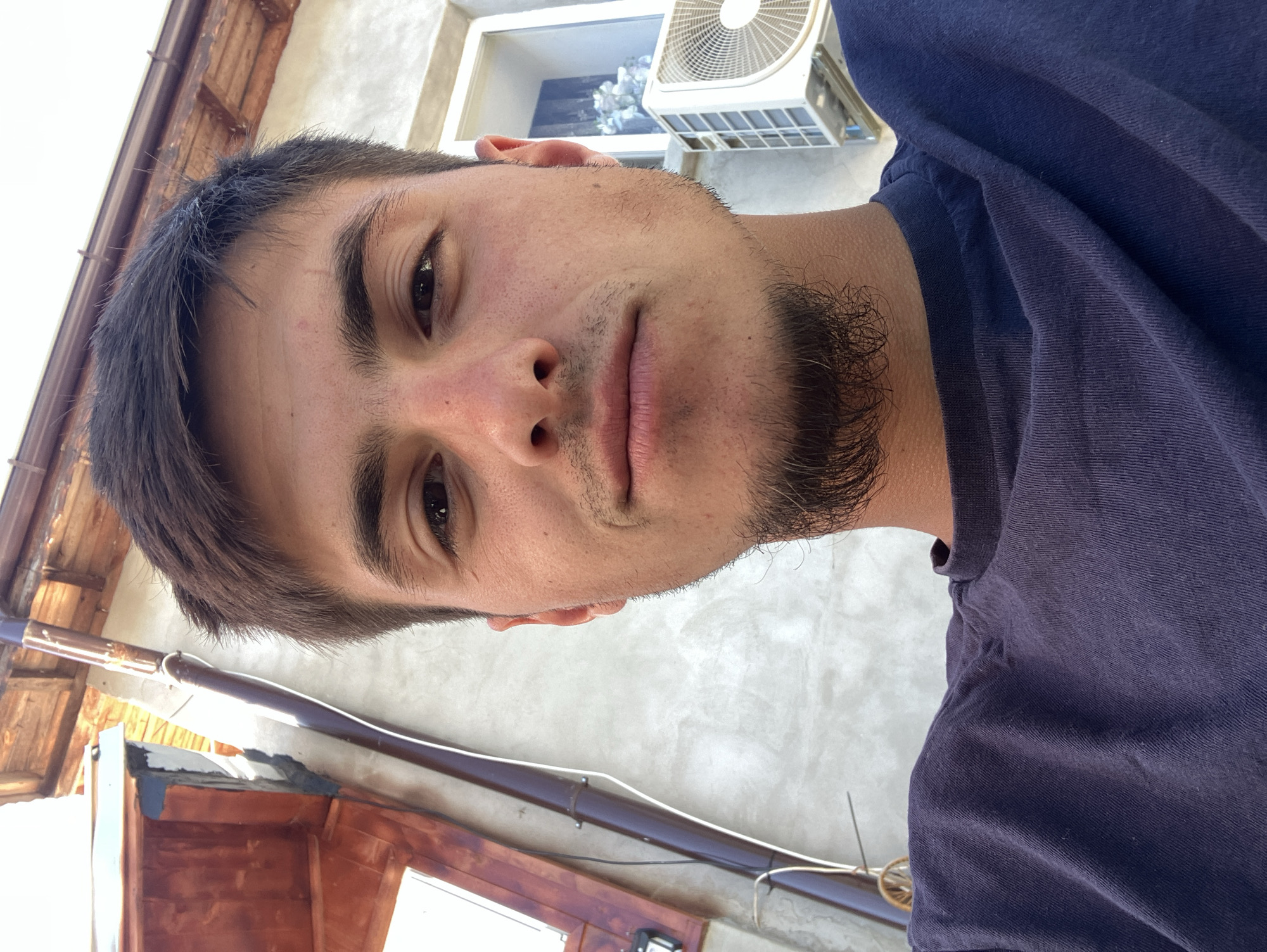A close-up, upward-angled photograph shows a young adult male with brown hair and eyes, light skin, and a sparse beard and mustache. He is wearing a dark blue t-shirt. Behind him and slightly above are parts of a building exterior: a window with a white air conditioning unit, a roof overhang with wooden elements, and a light-colored wall.
The city is mentioned, but there is no visual information to support the claim that the picture was taken there.