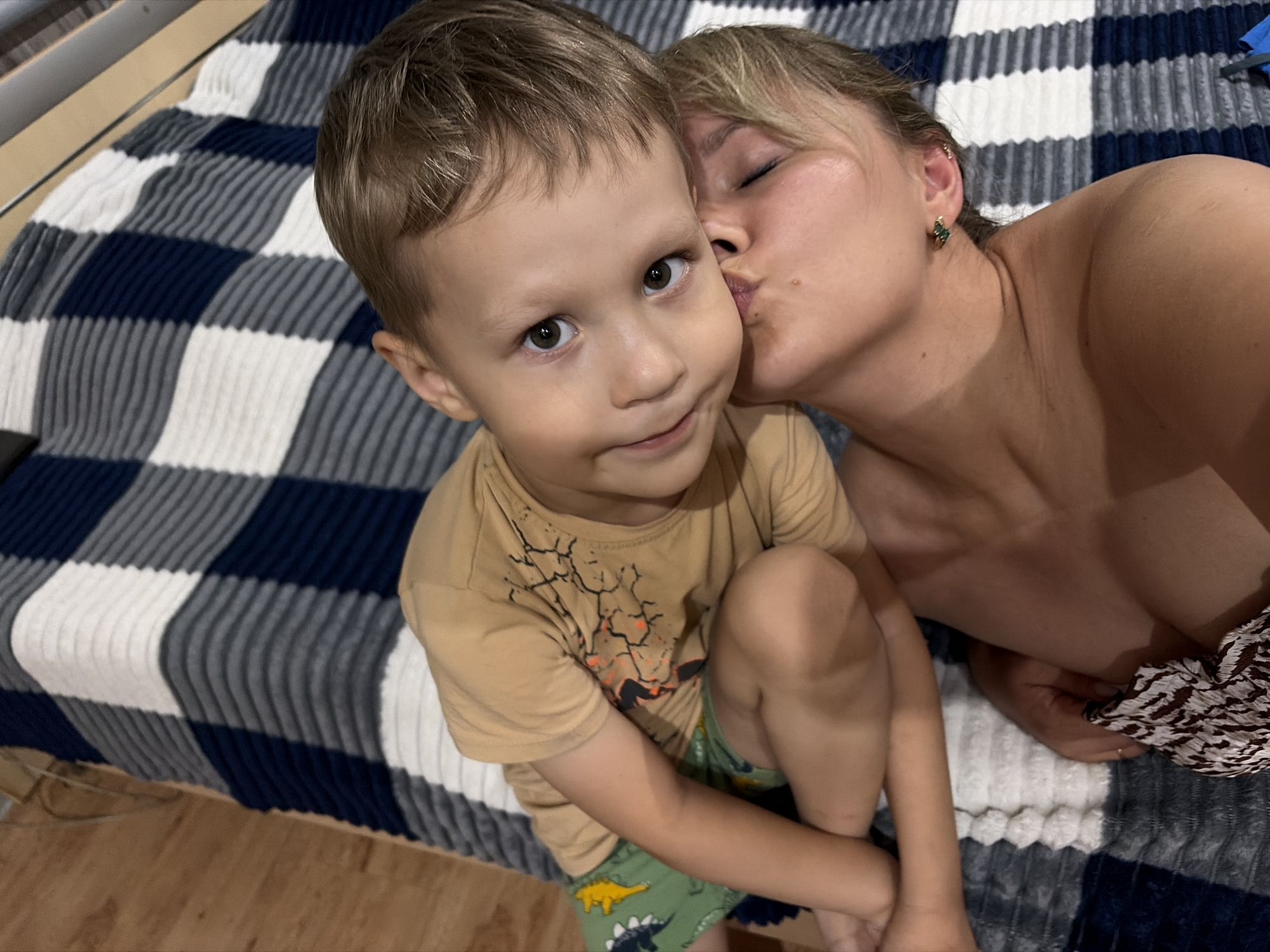 In this close-up selfie, a woman is kissing a young boy on the cheek. The boy is looking directly at the camera with a gentle smile. He is wearing a tan t-shirt with a graphic design and green shorts with a dinosaur print. The woman has blonde hair pulled back and is wearing a green earring. They are both lying on a bed with a checkered blanket featuring navy blue, gray, and white stripes. The background includes a wooden floor and what appears to be a bed frame. The overall mood is intimate and affectionate, suggesting a warm family moment. The lighting suggests it could be daytime.