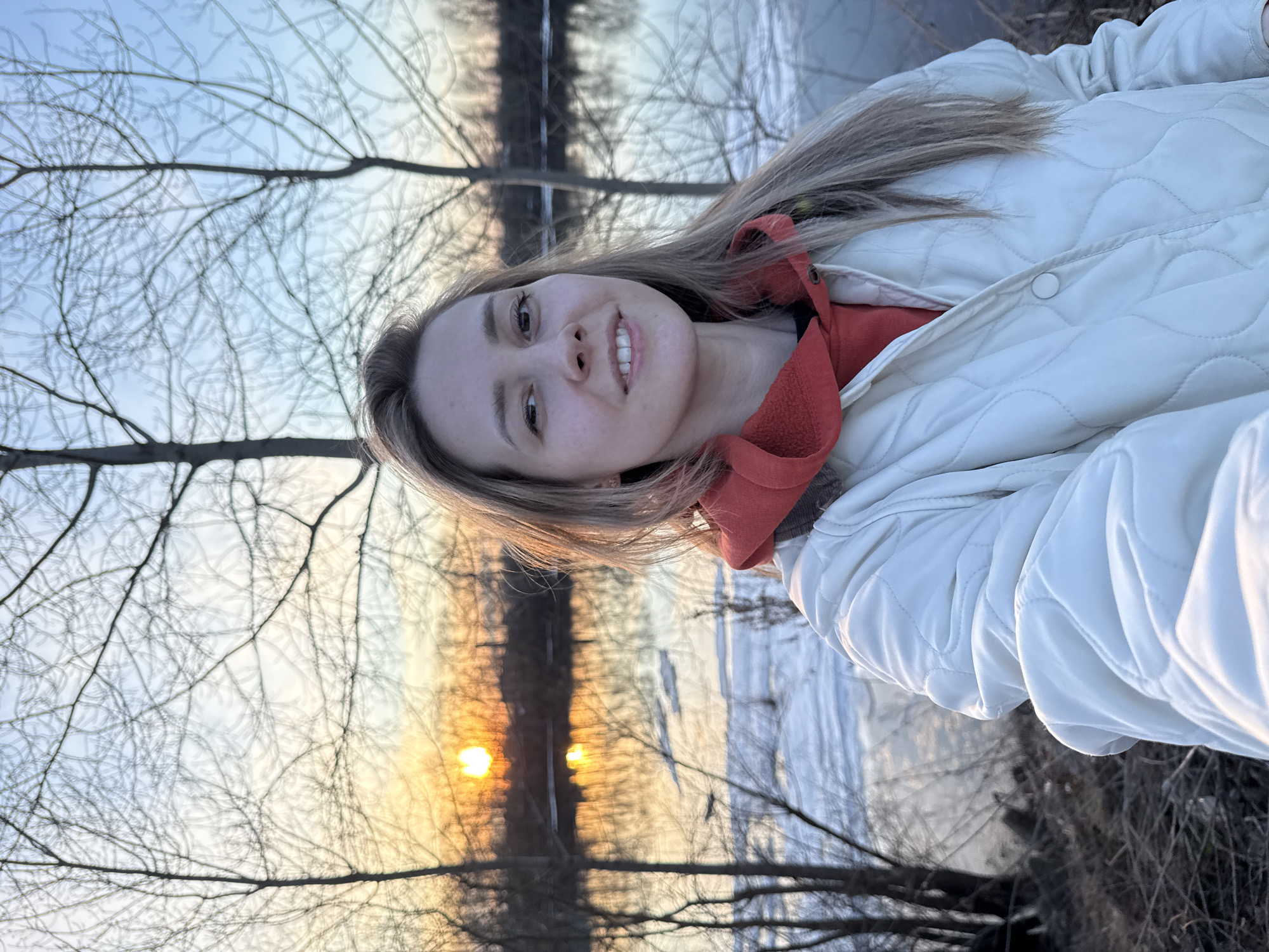 A young woman poses for a selfie outdoors. She has fair skin, light brown hair, and is smiling. She is wearing a white quilted jacket over a red hooded sweatshirt. Behind her, the sun is setting, casting a warm glow through bare tree branches. In the background, there appears to be a body of water, possibly a river or lake. Given the context of Chaikovsky, Russia, it could be the Kama River or one of the nearby lakes or ponds, but without further visual cues, this cannot be definitively confirmed.