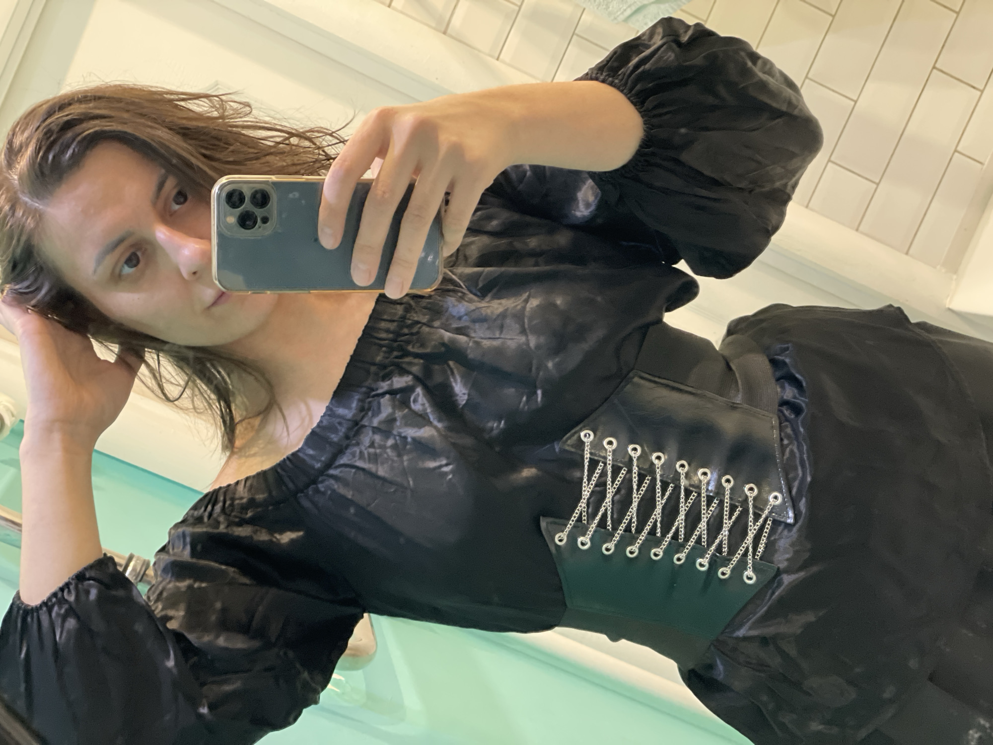 A woman takes a selfie in what appears to be a bathroom. She is holding a smartphone in her right hand. She is wearing a black, off-the-shoulder dress with puffy sleeves. The dress appears to have a corset-style detail with a chain design on the torso. Her hair is damp and parted to the side. The background shows a white-tiled wall with a blue-green surface below, likely part of a sink or shower area. The location appears to be an indoor setting in an Unknown city.
