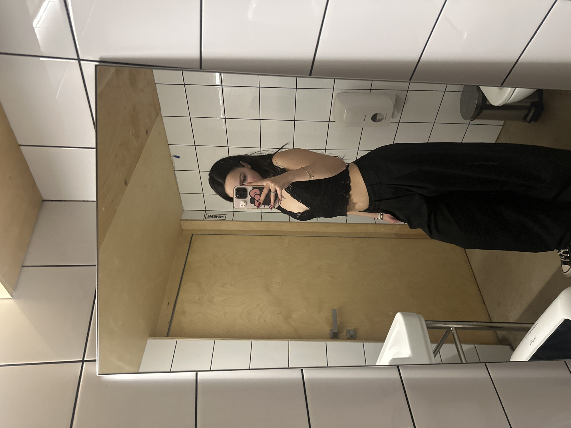 A person is taking a selfie in a public bathroom. The bathroom has white tile walls and a light wood door. There is a white sink and a toilet visible. The person is wearing a black lace top and black pants. The phone case is pink with cartoon characters. There is no information about the location in the city of Yerevan, Armenia.
