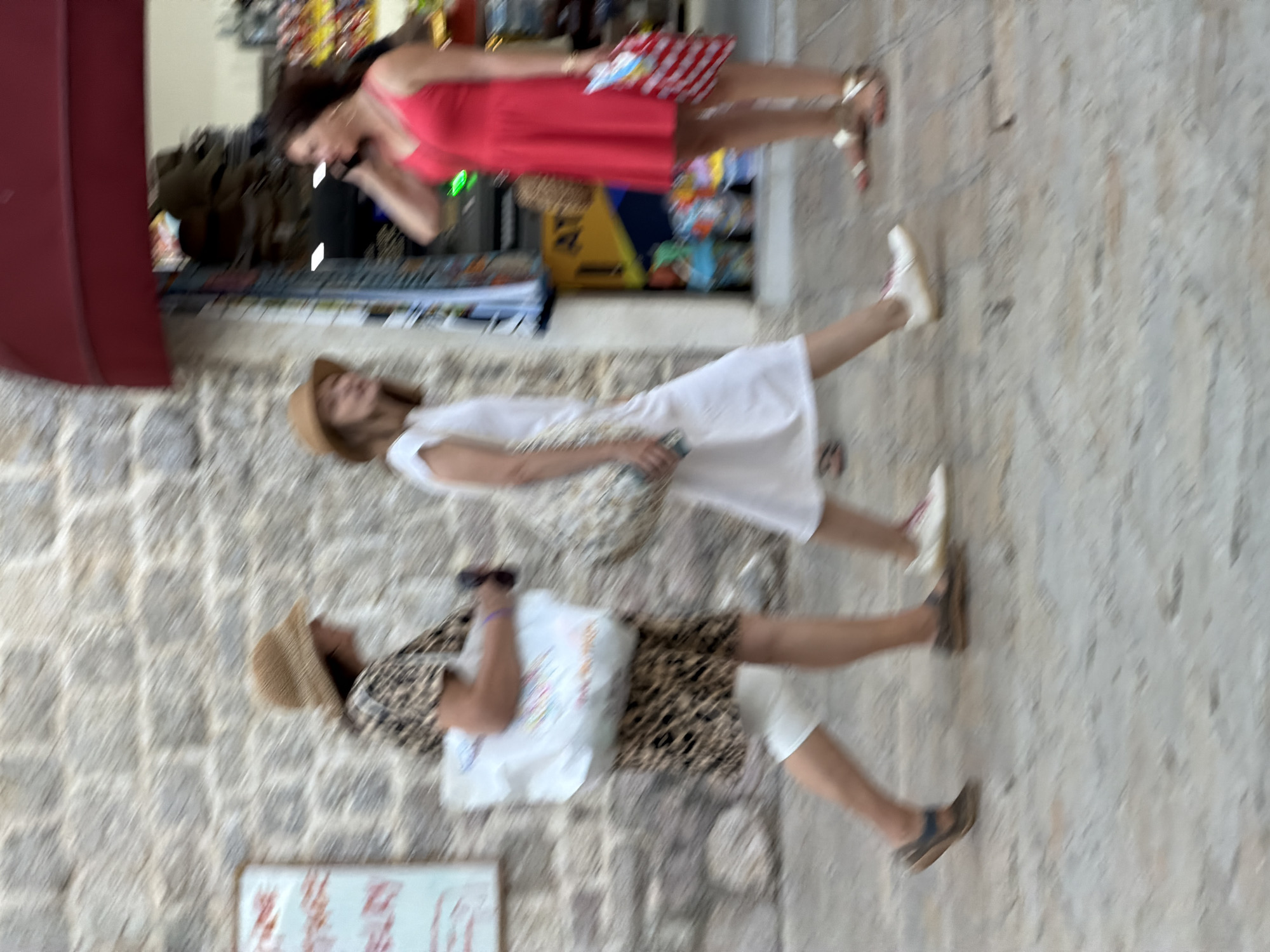 The image depicts a street scene in Budva, Montenegro, blurred due to motion. Three individuals are visible, all women, walking along a paved walkway adjacent to a stone building and a shop with a red awning.
The woman on the left, wearing a straw hat and a patterned dress, carries a large white tote bag. The woman in the middle, also wearing a straw hat, is dressed in a white dress and appears to be walking ahead of the first woman. The woman on the right, wearing a bright red dress, is talking on her phone and holding a striped shopping bag.
The environment suggests a warm climate, with people dressed in light clothing. The shop in the background displays various merchandise. The stone building on the left has a sign with some text, partially obscured. The ground is a textured pavement, typical of older European city streets. The overall impression is of a casual, bustling urban environment.