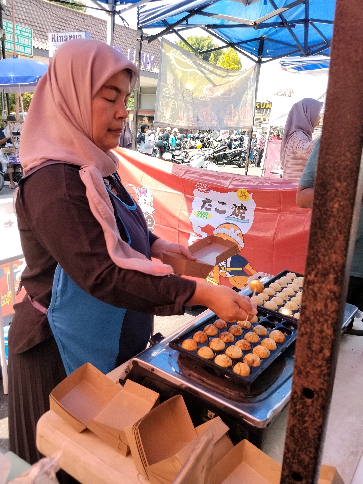 In an outdoor market stall, likely in Depok, Indonesia, a woman wearing a pink hijab and an apron is preparing takoyaki. She is holding a small wooden skewer, turning a round, golden-brown takoyaki ball in a specialized cooking pan. The pan contains numerous similar takoyaki balls, some of which appear to be freshly cooked and golden, while others are still in the process of cooking. In front of the cooking station, several small, open brown cardboard food containers are stacked on a light-colored surface. Behind the woman, a red banner with Japanese text ("たこ焼" and "TAKO YAY!") and cartoon illustrations is displayed. The background shows a busy market scene with motorcycles, other stalls, and people. The overall atmosphere suggests a bustling street food vendor operation.