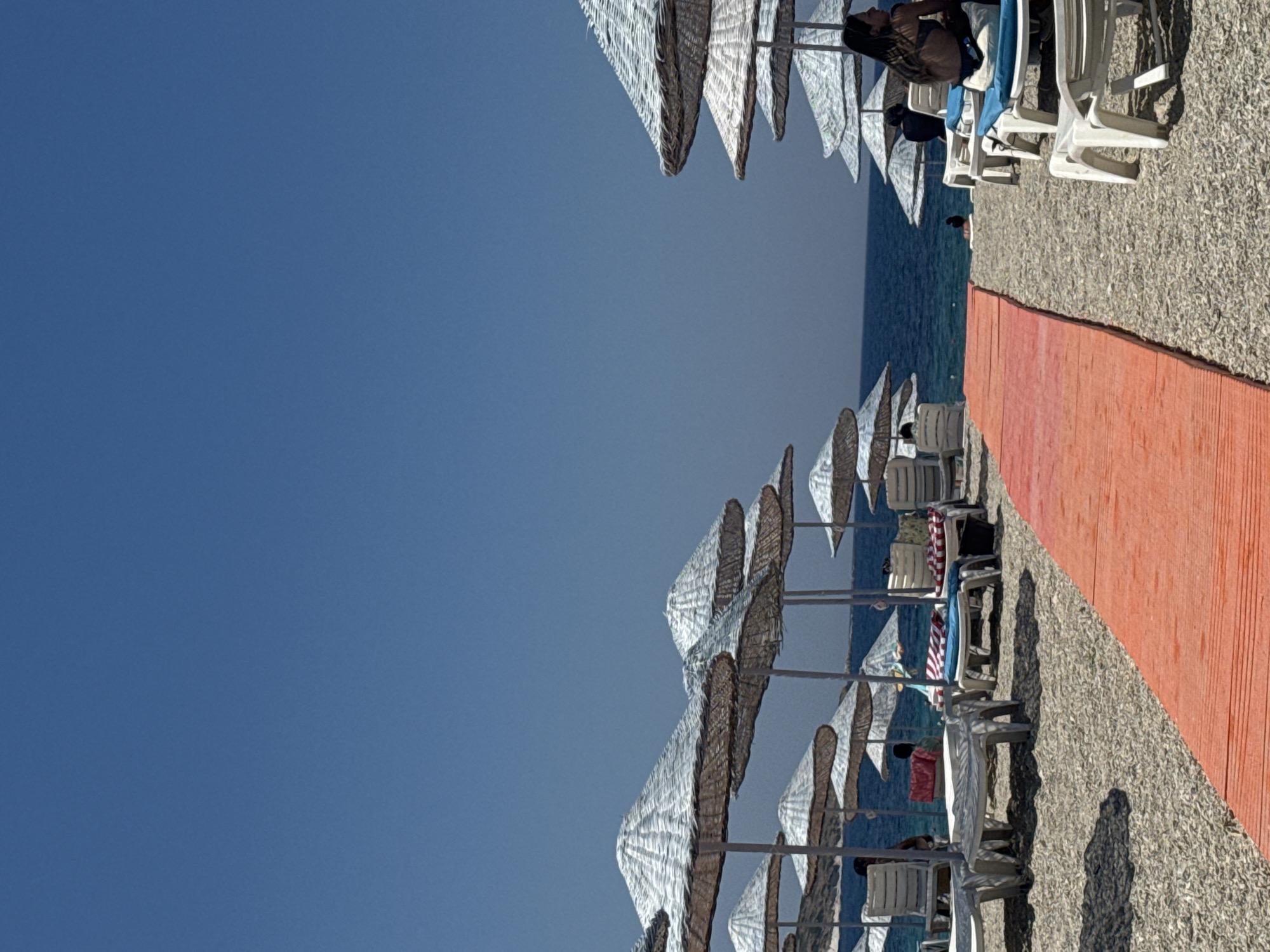 This is a vertical, eye-level shot of a beach in Foça, Turkey, under a clear, bright blue sky. In the foreground, a vibrantly colored orange walkway extends diagonally across the frame from the bottom right to the middle left. The walkway is made of what appears to be wooden planks. To the right of the walkway, several white plastic lounge chairs are visible on a pebbly beach, some with blue cushions. On the left side of the walkway, more lounge chairs are arranged, with some partially obscured by large, conical thatched umbrellas.
In the middle ground, the blue sea stretches towards the horizon. Beyond the beach, there are rows of thatched umbrellas providing shade. A few people are present on the beach, some sitting in lounge chairs or standing. On the far right, a woman with long dark hair is sitting on a lounge chair facing away from the camera, wearing a bikini. Further back, near the water's edge, a person is partially visible. The overall scene depicts a typical beach resort environment on a sunny day.