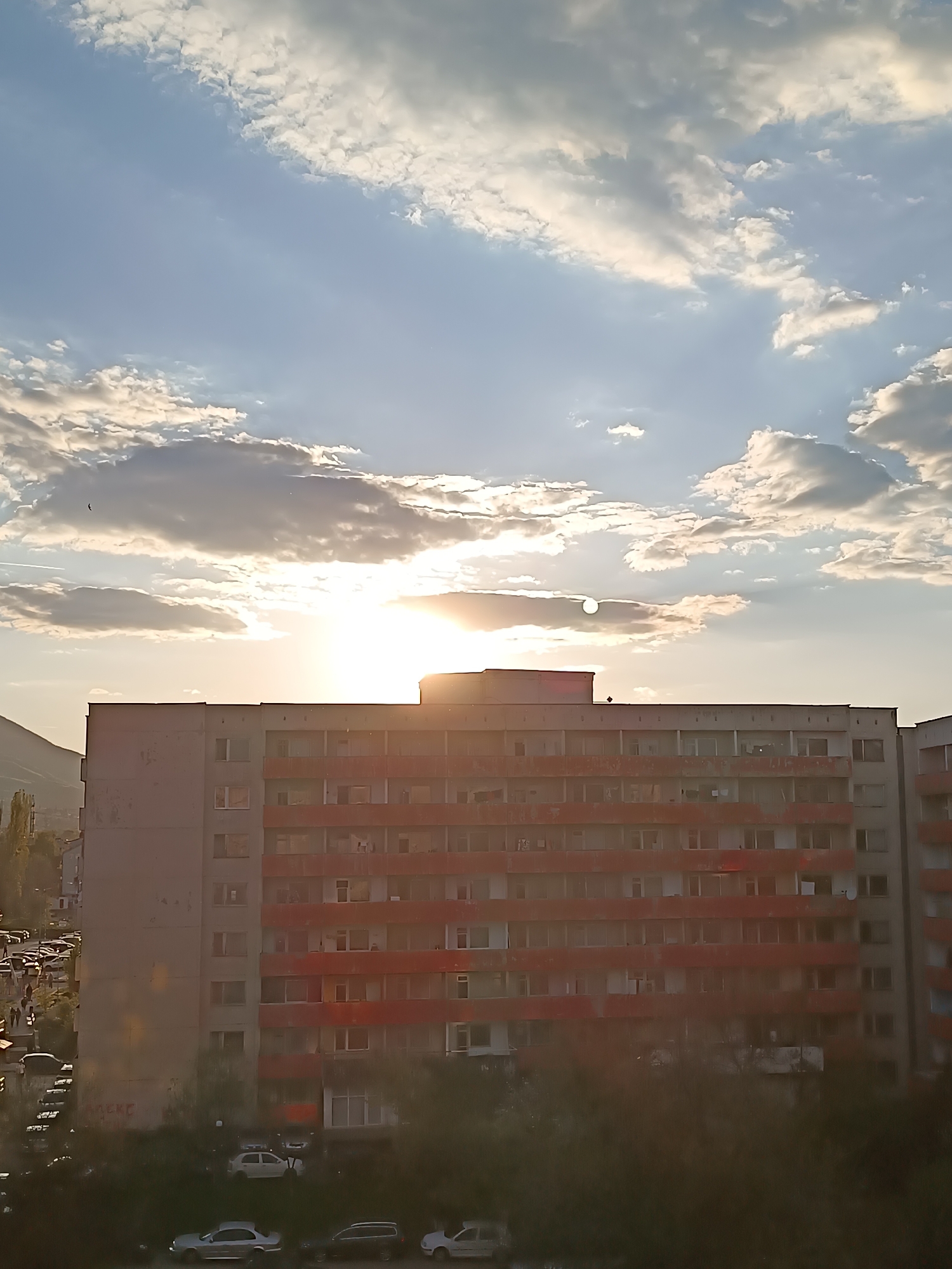 The image presents an urban residential scene captured during sunset or late afternoon, characterized by the low, bright sun. The central focus is a large, multi-story apartment building, typical of socialist-era panel block architecture. The building's facade is predominantly light grey, accented by prominent reddish-orange balconies or sections. Numerous windows are visible across its many floors, some revealing interior activity or curtains.
Above the building, the sky is partly cloudy, with white and light grey clouds scattered against a light blue expanse. The sun, a brilliant white orb, is positioned behind the building, casting a strong glare and warm light that highlights the cloud edges and creates a lens flare effect. To the far left, the silhouette of a mountain or large hill is faintly visible, suggesting the city's geographical context.
In the lower foreground and to the left of the building, there are multiple cars parked, along with green trees and vegetation. Small, indistinct figures, possibly people walking, can be seen near the parked vehicles on the left side of the frame. On the lower part of the building's facade, faded, light-colored text resembling graffiti is visible, appearing to read "RAKEC" or a similar sequence of letters. The overall scene conveys an everyday moment in a densely populated urban environment, consistent with residential areas in Sofia, Bulgaria, where such architecture is common.