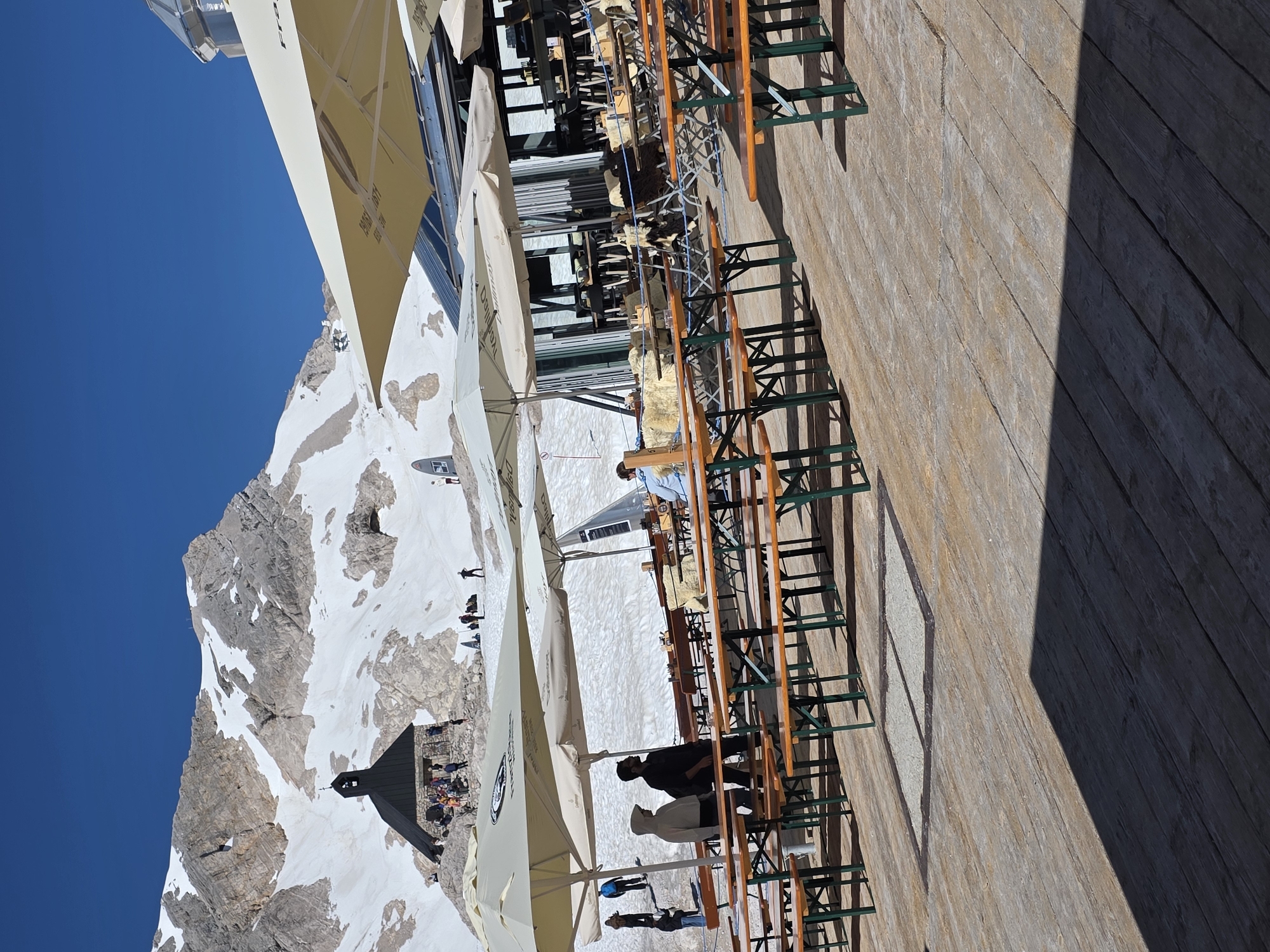 The image captures a sunny day scene at a mountaintop location, likely the Zugspitze, near Garmisch-Partenkirchen, Germany. In the background, a snow-capped mountain range dominates the horizon under a clear blue sky, with a small dark-roofed building with a bell tower nestled on the mountainside, surrounded by a scattering of people. In the foreground, a wooden deck is set up with numerous picnic tables and benches, most of which are empty. Several large, light-colored umbrellas are deployed over parts of the deck, providing shade. A few individuals are visible on the deck, some standing near the tables and others in the mid-ground near the building, appearing to be tourists or patrons of an outdoor cafe. The overall impression is one of a popular tourist destination offering scenic views and outdoor dining opportunities.