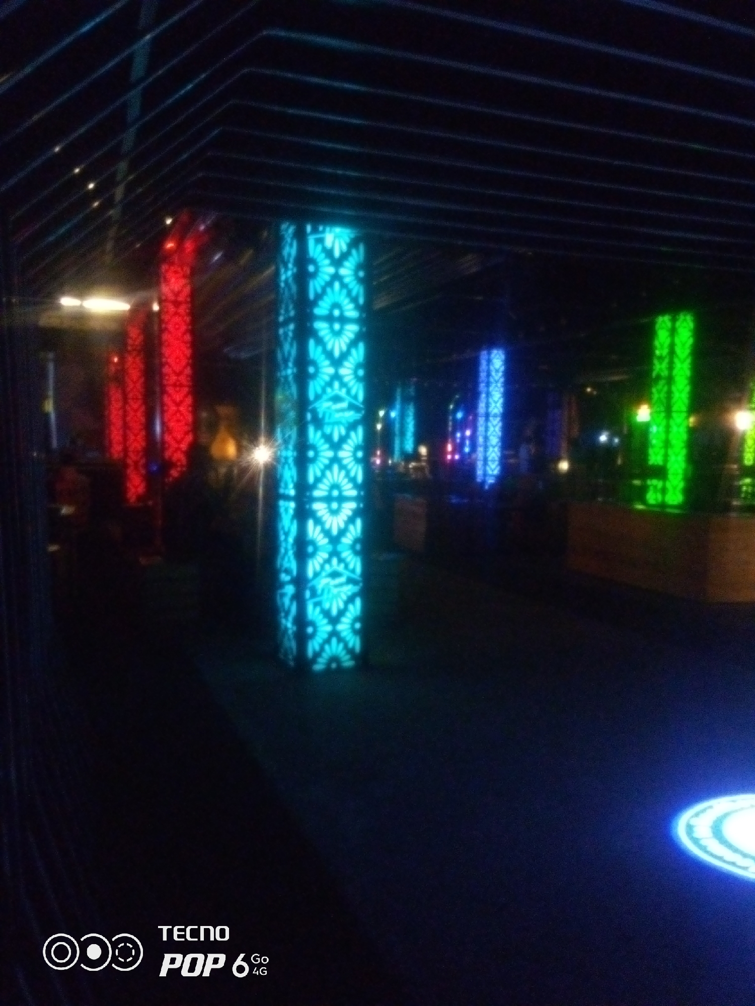 In Howayi, Nigeria, this image captures an indoor setting with an emphasis on illuminated columns. Multiple tall, rectangular columns are prominently featured, each emitting a distinct color of light. Closest to the viewer is a column glowing bright cyan, patterned with a floral or star-like design. To its left, a column is lit in vibrant red, displaying a similar decorative pattern. Further in the background, other columns glow blue and green, also featuring the patterned illumination. The environment appears to be a dimly lit space, possibly a lounge, club, or restaurant, with a dark floor and a ceiling featuring horizontal dark lines. A bright light source, possibly a camera flash, creates a strong glare in the middle ground. The bottom right corner of the image shows a circular, glowing element. In the bottom left, the "TECNO POP 6 4G" logo is visible.