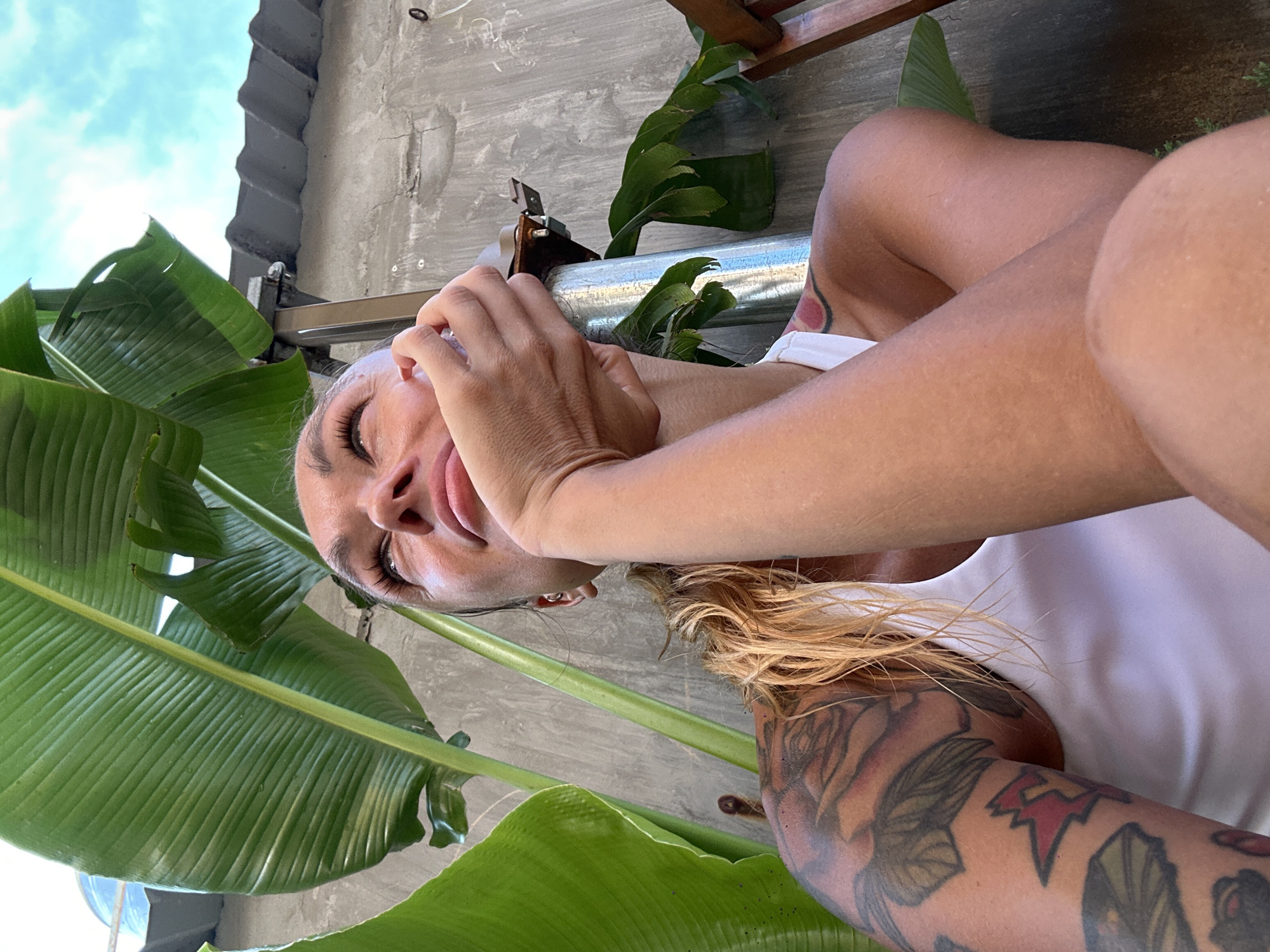 A woman with blonde hair and tattoos on her arm is sitting outside near large green leaves. She is looking up at the sky and resting her head on her hand. The background is a concrete wall and a tiled roof. The scene appears to be in a residential area in Phu Thuy Ward, Vietnam, likely a courtyard or backyard.