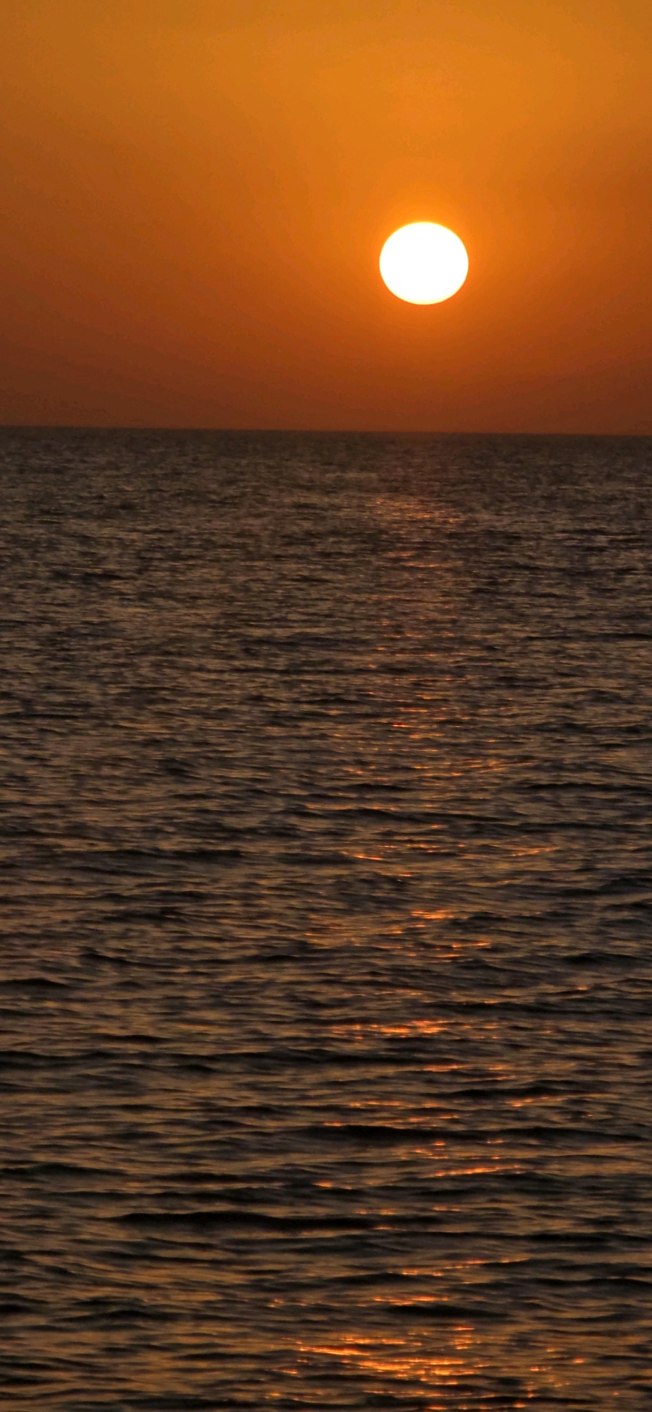The image shows a sunset over a body of water. The sun, a bright white orb, is positioned in the upper right quadrant of the frame, casting a warm orange glow across the sky. The horizon line where the sky meets the water is clearly defined. The water's surface is textured with ripples, and reflections of the sunset create streaks of orange light across it. The lower portion of the image is dominated by the dark, rippling water. There are no people or man-made structures visible in the scene, suggesting a natural, open environment. The provided location of Basbeshik, Kyrgyzstan, is not visually verifiable from the image itself, as there are no identifying landmarks.