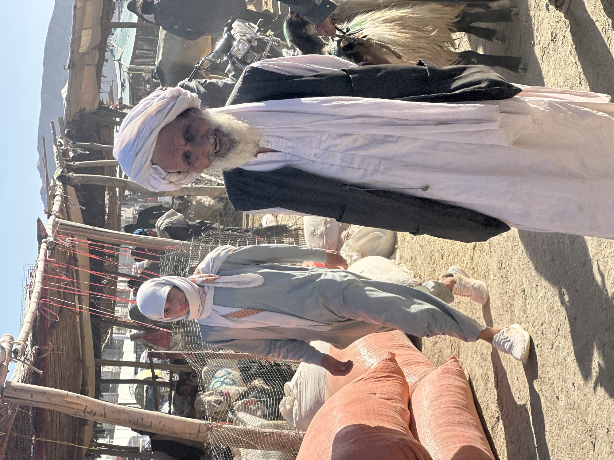 In Quetta City Tehsil, Pakistan, an older man with a white turban, beard, white shirt, and black vest stands with a slight smile. Beside him stands a younger person in a light-colored robe and head covering. They are amidst a market scene with people, animals, motorcycles, and sacks. The setting appears to be outdoors with a makeshift structure visible in the background.