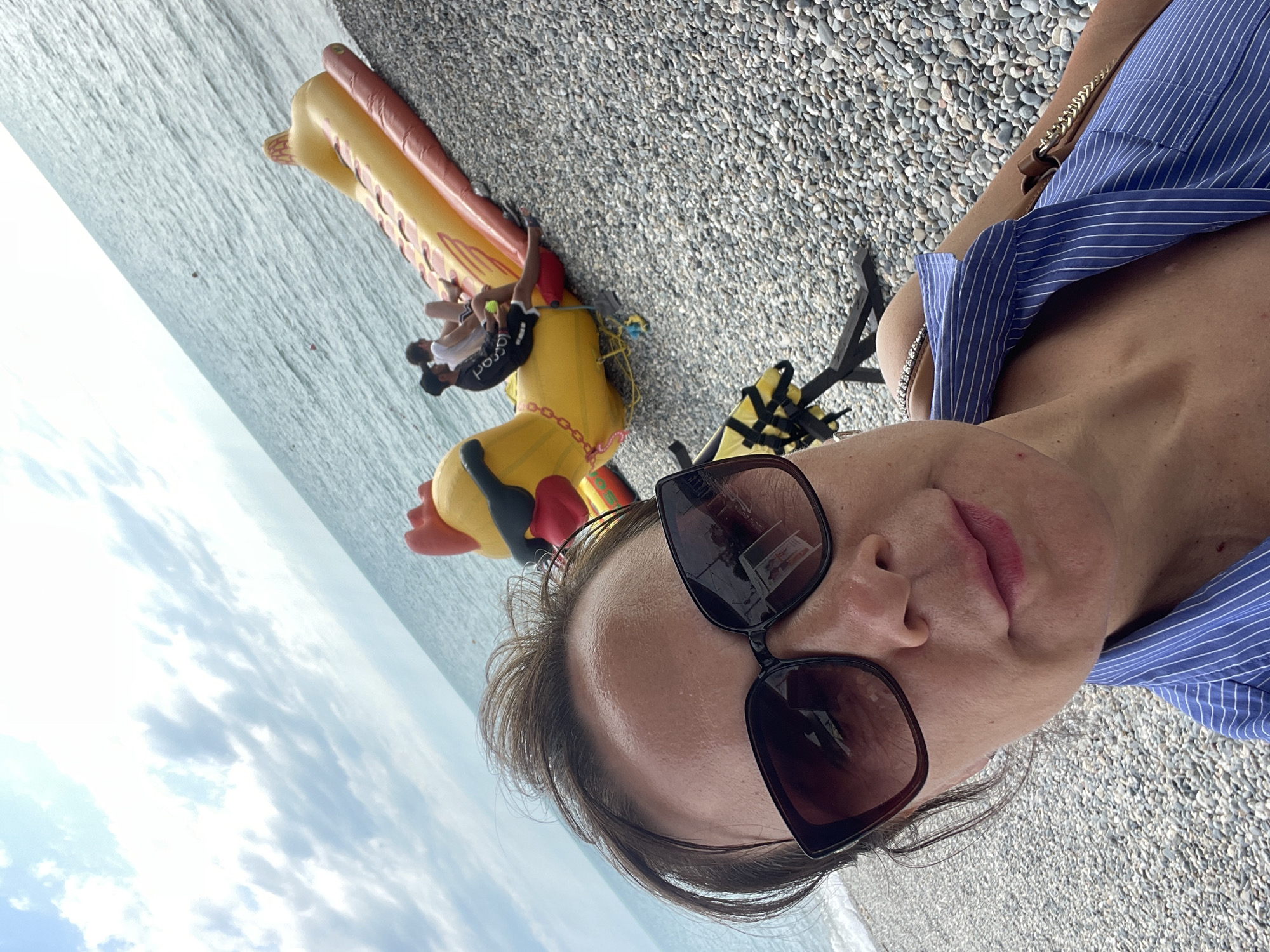 The image depicts a close-up selfie taken outdoors on a pebble beach, with the sea visible in the background and a cloudy sky above. The primary subject is a woman wearing sunglasses and a blue and white striped shirt. Her face is the central focus, with her lips slightly parted and her hair falling around her face.
To the left of the woman, a large, inflatable yellow duck-shaped pool float is partially visible, lying on the beach. Two young children, a boy and a girl, are interacting with the inflatable toy. The boy, wearing a white t-shirt and dark shorts, is seated on the float, while the girl, in a white top and dark shorts, is also on or near the float.
The setting suggests a sunny day, likely during the daytime given the brightness and the clouds. The presence of the inflatable and the beach strongly indicates a recreational activity, likely a family outing to the seaside in Sochi, Russia. The overall mood appears relaxed and casual. No discernible text is visible in the image.
