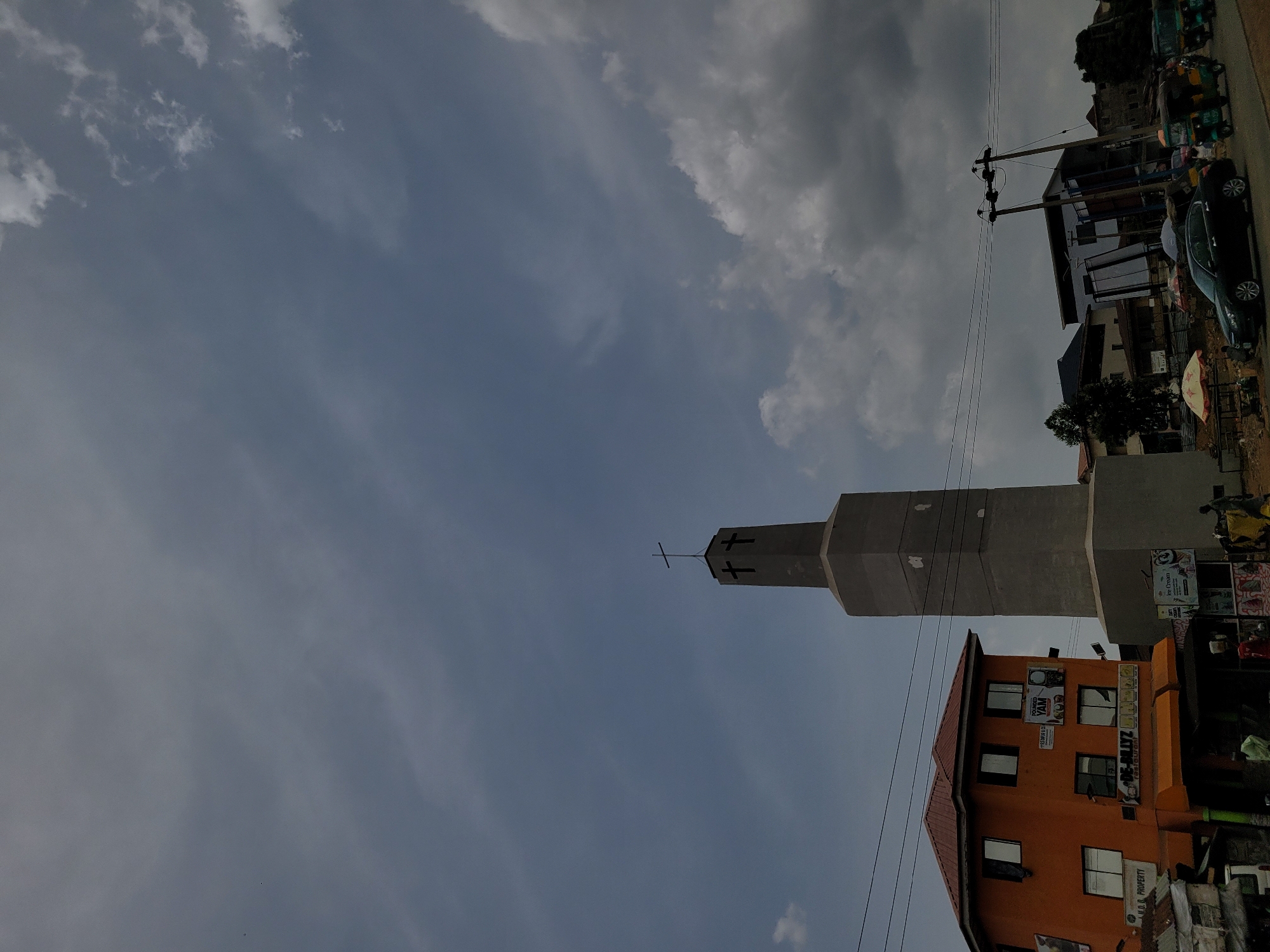 The image shows a church bell tower in Jos, Nigeria. The tower is made of light gray concrete and has a cross at the top. Two black crosses are visible on the side of the tower. The tower is surrounded by other buildings, including an orange building with windows. Power lines are visible. The sky is cloudy. Vehicles and people are visible on the ground.