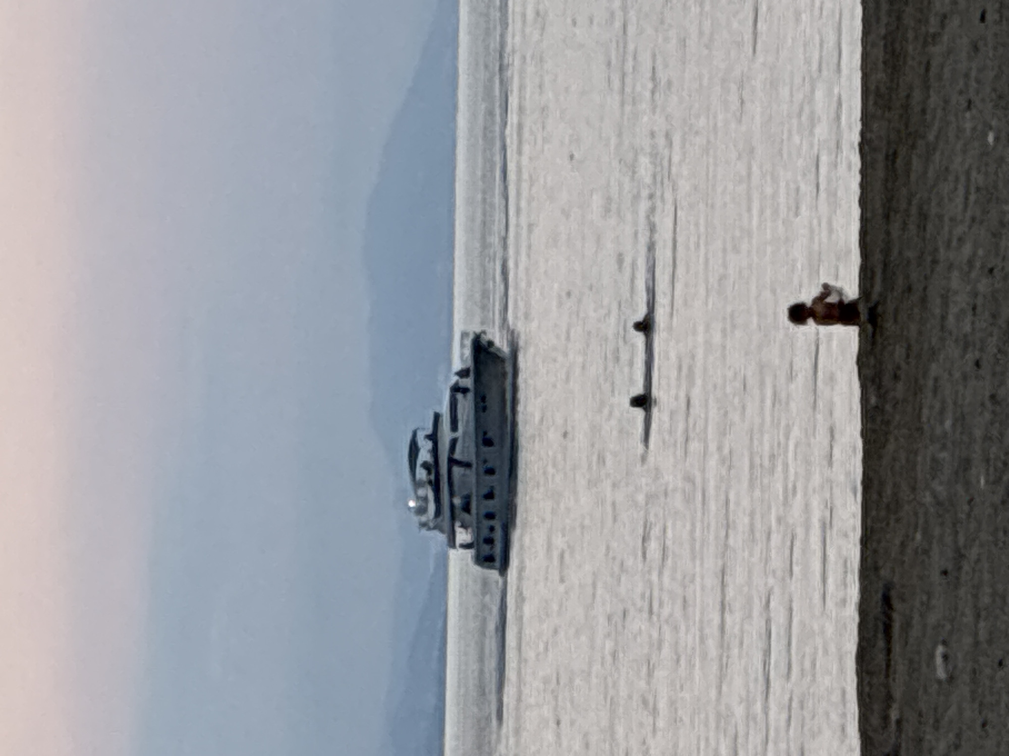 The provided image, which appears to be rotated 90 degrees clockwise from its natural landscape orientation, depicts a calm coastal scene, likely in Alanya, Turkey. The majority of the frame is dominated by a placid body of water, possibly the Mediterranean Sea, stretching towards a hazy horizon. The water's surface is subtly textured by gentle ripples and reflects the soft daylight.
In the middle-left of the water, a substantial multi-decked yacht or boat is visible, featuring several levels, windows, and what appears to be an antenna or mast on its top deck. To the far right, a dark, textured structure, resembling a pier or the immediate shoreline, forms a vertical boundary against the water. A small, indistinct figure, possibly a child, stands or sits at the water's edge on this dark structure, facing out towards the sea.
Between the large boat and the figure, two smaller, dark, elongated objects float on the water, their exact nature unclear but possibly buoys or parts of watercraft. In the distant background, a faint, light blue sky meets a soft, undulating line of what appears to be mountains or distant landmass. The scene is bathed in a gentle, diffused light, suggesting either early morning or late afternoon on a clear, calm day. No discernible text is visible within the image. The overall impression is one of peaceful seaside leisure.