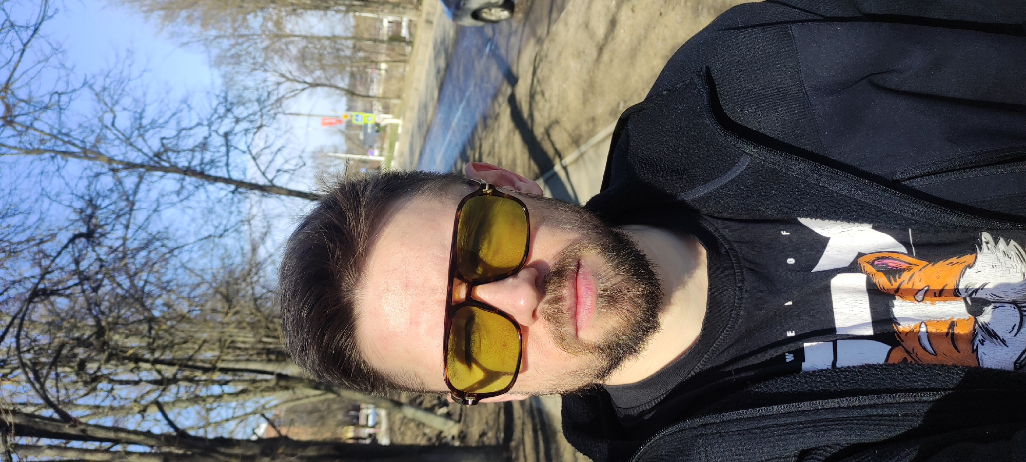 The photograph captures a close-up, angled shot of a man wearing tinted sunglasses, a black hoodie, and a black t-shirt with a graphic featuring a stylized fox and the words "WE LOVE OF" on it. The environment suggests an outdoor location, possibly a street or park, given the presence of trees and visible infrastructure in the background. The lighting indicates daytime, with strong sunlight casting shadows. The location is the city of Fryazino, Russia. The image is oriented vertically.