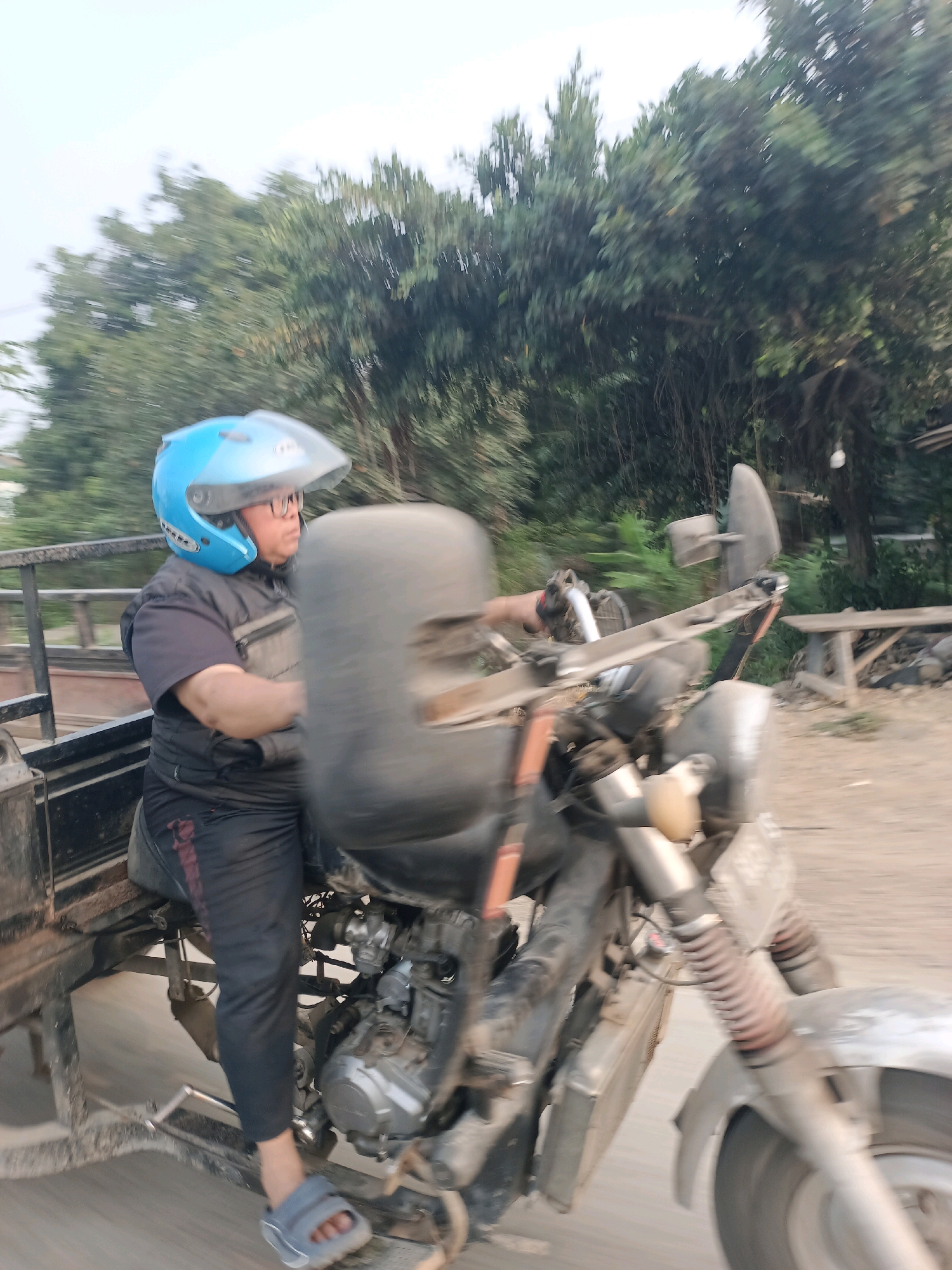A person wearing a blue helmet and glasses is riding a motorized tricycle on a road lined with trees. The person is wearing a black vest over a black shirt and black pants with a red stripe. The tricycle has a large black seat in the middle and a metal cargo bed in the back. The scene is blurred due to motion. The location is Subang, Indonesia.