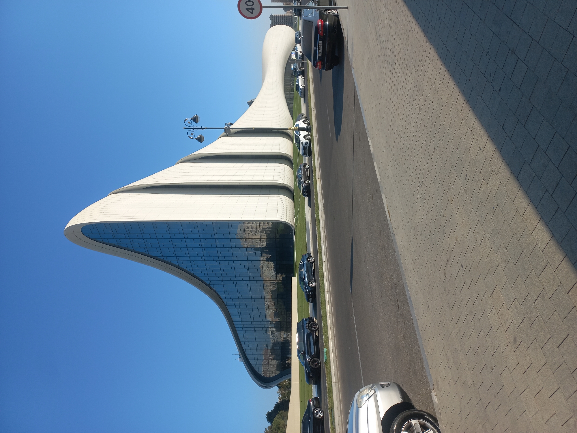 The Heydar Aliyev Center, a distinctive architectural landmark in Baku, Azerbaijan, is showcased on a clear, sunny day. The building's fluid, wave-like design, characterized by its white, undulating exterior and extensive glass facade, dominates the background. The facade reflects the blue sky and surrounding buildings. In the foreground, a paved pedestrian walkway lines a multi-lane road where several vehicles are visible, including black sedans and a dark-colored SUV. A speed limit sign indicating "40" is posted on a pole at the right side of the image. The scene captures a typical urban environment with a focus on the iconic structure.