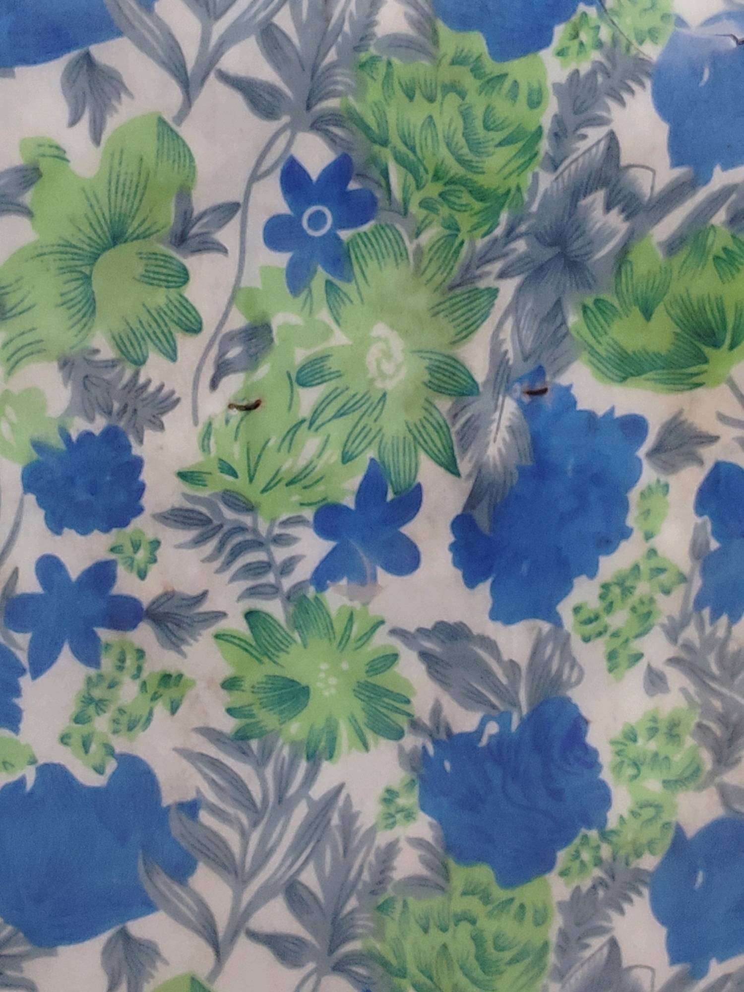 The image displays a close-up, top-down view of a floral fabric with a white background. The pattern features a mix of blue and green flowers, along with grey foliage. The flowers vary in size and style, some appearing as simple five-petal blossoms in blue, while others are more intricate, with layered green petals. The grey elements resemble leaves and stems, creating a naturalistic yet stylized design. The texture of the fabric is visible, suggesting it might be cotton or a similar material. The lighting is diffused, and there are no shadows, indicating an indoor setting or an overcast day. The image is static and does not depict any specific location within Jamshedpur, India, or any people or actions.