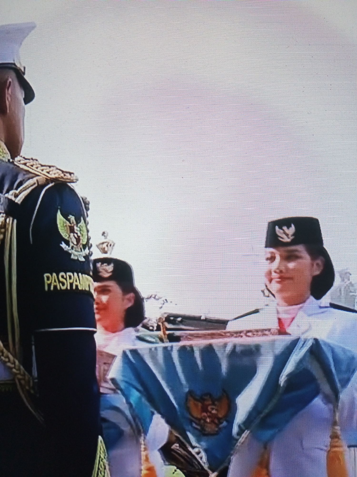 The image depicts a formal ceremony in Banyudono, Indonesia, likely at the Tri Satya Building. In the foreground, a man in a dark uniform with gold trim and a white hat stands to the left, his back partially to the camera. His uniform features a crest on the left sleeve and the word "Paspam" embroidered in gold on the back. To the right, two women in white uniforms with black caps stand. The woman on the right, closer to the camera, is looking to her left. Both women appear to be participating in the ceremony. Between them, a light blue cloth is draped over an object, adorned with a golden crest. The background is a bright, overcast sky.