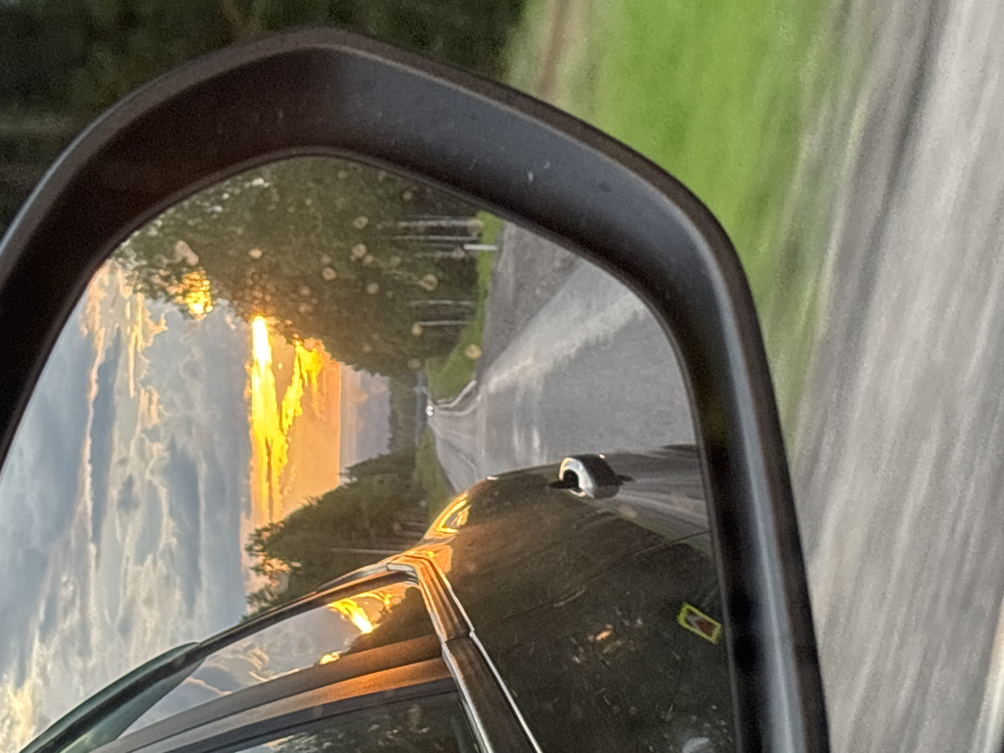 The image captures a scenic view of a road stretching into the distance, reflected in a car's side-view mirror. The sun is setting, casting a warm golden glow across the sky and illuminating the clouds with hues of orange and yellow. Trees line the sides of the road, their reflections visible in the mirror alongside the road and the sky. The road itself appears to be paved with gravel and asphalt, and a few distant vehicles can be seen. The car, partially visible in the foreground, is dark in color and has a side-view mirror with some water droplets on it. The environment suggests a rural or semi-rural setting, possibly in Ivakinskii Kar'er, Russia, given the presence of trees and a road that could be found in such a location. The overall impression is one of a peaceful journey during sunset.