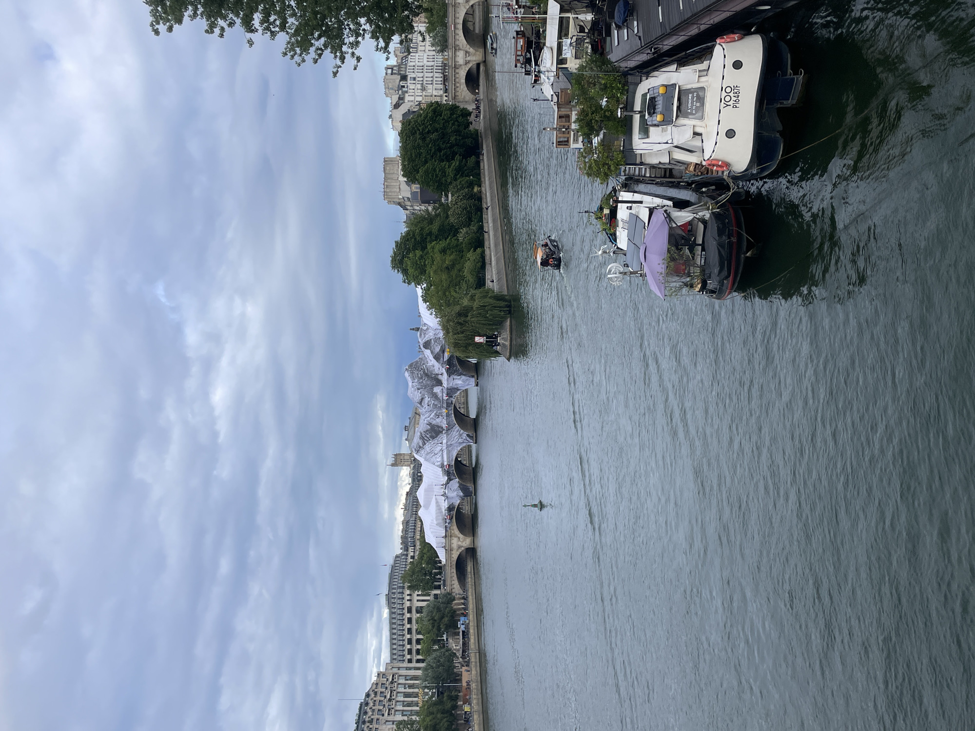 The image depicts a view of the River Seine in Paris, France, with a partially wrapped Pont Neuf bridge in the background. The bridge is draped in white fabric, giving it an abstract, sculptural appearance. On the right side of the foreground, several houseboats are moored, one of which has "YOO P16487F" and "À VENDRE" written on its stern. The river is calm with gentle ripples, and a small boat is moving in the distance. The sky is overcast with diffused light, suggesting a cloudy day. Trees and buildings line the riverbanks, contributing to the urban landscape of Paris.