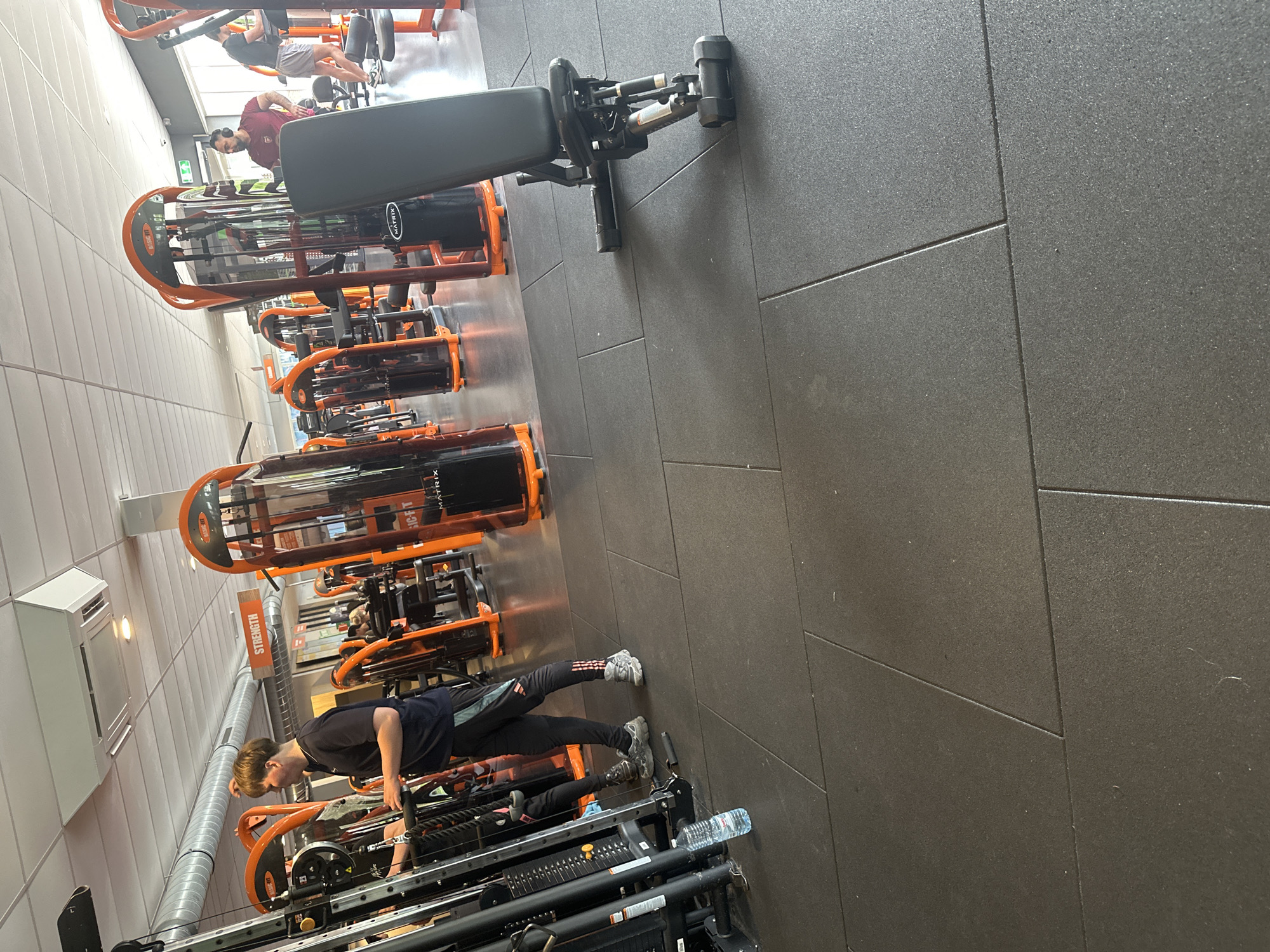 The scene depicts the interior of a Basic-Fit gym located in Schagen, Netherlands. The environment features a grey rubber floor composed of large interlocking tiles, a white acoustic panel ceiling with recessed lighting, and exposed silver air ducts. Numerous orange and black gym machines, predominantly branded "BASIC-FIT" and "MATRIX," are arranged throughout the space. An orange sign labeled "STRENGTH" is visible in the mid-ground.
In the left foreground, a young male with light brown hair, wearing a dark blue short-sleeved shirt and dark athletic pants with light grey stripes and sneakers, stands facing a multi-functional cable machine. He is actively engaged with the machine, with his hands positioned on its components. A clear plastic water bottle with a blue label rests on the floor at the base of this machine.
In the mid-ground to the right, a black adjustable weight bench is positioned at an angle, facing away from the viewer. Further back, a man with dark hair and a beard, wearing a maroon t-shirt and headphones, stands near other gym equipment, looking towards the right side of the frame. In the background on the far right, another male figure, dressed in a dark t-shirt and light shorts, is visible near what appears to be a cardio machine. Several other indistinct figures are present in the deeper background, utilizing various pieces of gym equipment. Natural light enters the gym from large windows on the right side of the frame.