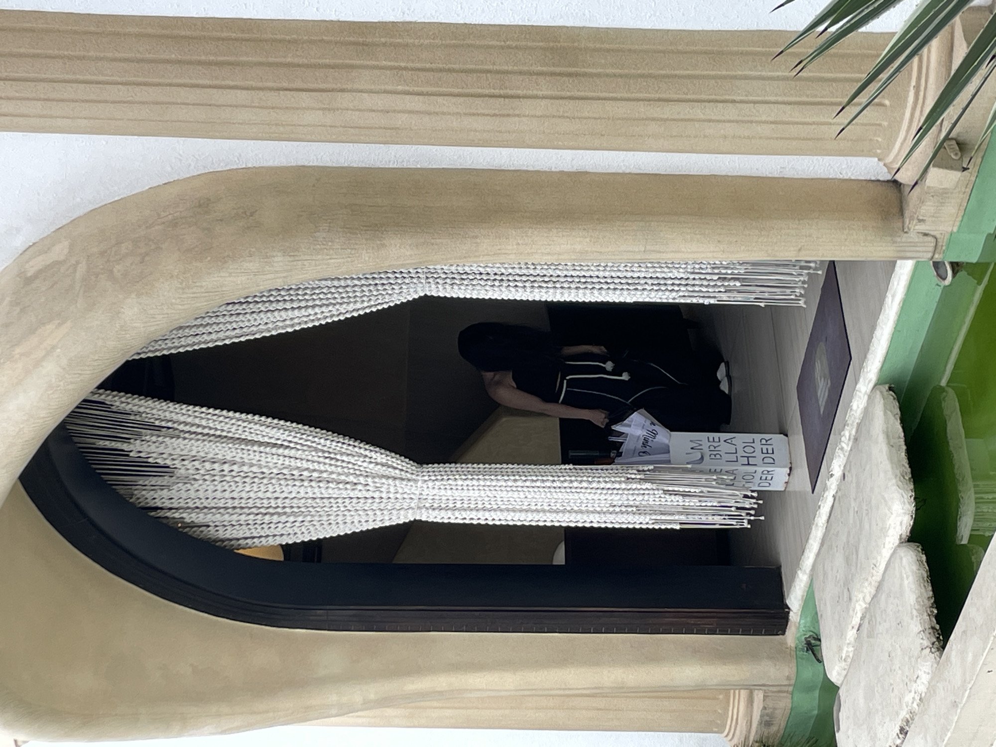 The image depicts a woman wearing a black strapless top and black pants with white drawstrings, carrying a white bag with black text. She is walking through a doorway draped with white beaded curtains. The doorway is framed by large, beige, curved architectural elements that resemble carved stone or stucco. To the right of the doorway, a fluted beige column stands against a white wall. In the foreground, a shallow pool of green water is visible, with two flat, light-colored stones forming a path across it. The environment suggests an entrance to a building or establishment, possibly a resort or a unique retail space. The provided image does not contain enough visual information to pinpoint a specific location within Da Lat, Vietnam.