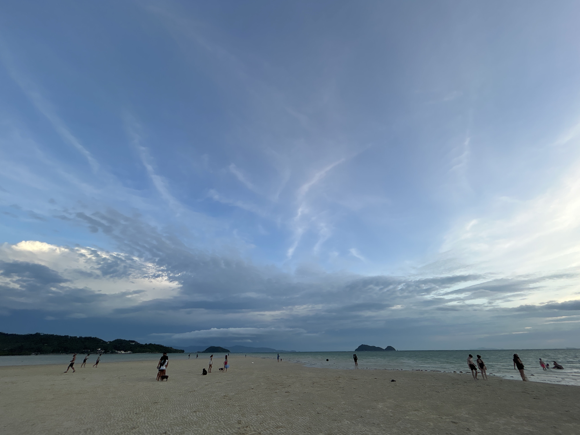 This image depicts a serene beach scene in Ko Pha-ngan District, Thailand, likely during the late afternoon or early evening given the soft, diffused light and the color of the sky. The sky is a prominent feature, filled with streaks of wispy clouds against a backdrop of pale blue, with some darker, more dramatic cloud formations visible on the horizon, suggesting a possible impending change in weather.
The foreground is a wide expanse of sandy beach, marked with rippled patterns from the receding tide or wind. Scattered across the beach are several people, appearing as small figures against the vastness of the setting. Some are walking along the shoreline, while others seem to be gathered in small groups, possibly chatting or observing the surroundings. A few individuals are closer to the water's edge, and at least one person is partially submerged in the sea, indicating a casual beach day activity. Two dogs are also present on the sand.
In the middle ground, the turquoise ocean stretches out, with gentle waves lapping at the shore. The horizon is dotted with several small, verdant islands, their silhouettes softened by the distance and atmospheric conditions. The overall mood of the image is tranquil and peaceful, capturing a moment of relaxation and natural beauty. There is no visible text in the image.