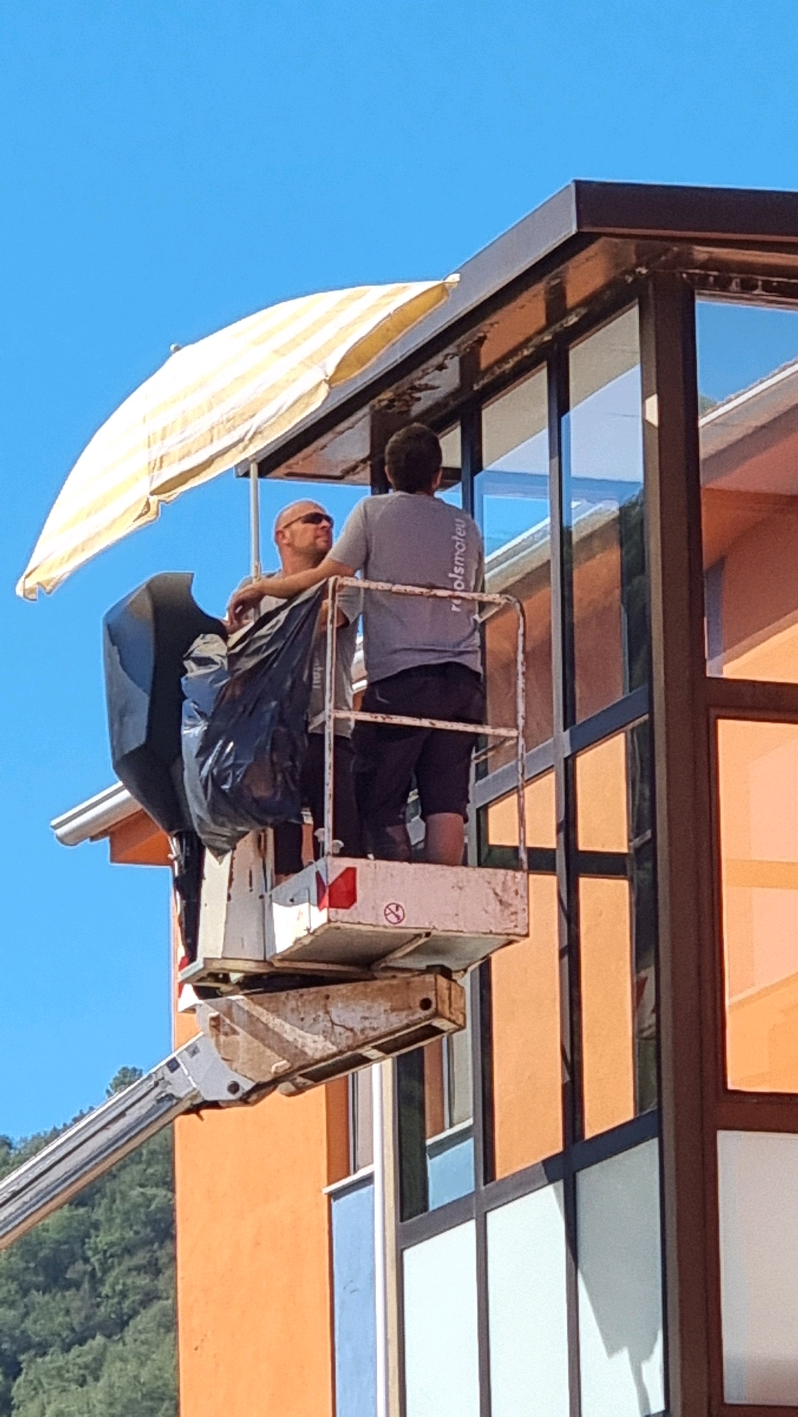 Two men are standing in the basket of a cherry picker, elevated against a bright blue sky. One man, wearing sunglasses and a grey t-shirt, is facing the other man, who is wearing a grey t-shirt with text and dark shorts. A yellow and white striped umbrella is positioned above and behind them, providing shade. A large black garbage bag is secured within the basket. The men appear to be working on the exterior of a modern building with large windows and an orange facade. The scene is outdoors, with a hint of green foliage visible in the background, suggesting a hillside or natural area. The building's architecture suggests it could be in a developed area, possibly a business or residential complex within the town of Sant Joan Les Fonts, Spain, given the context. The angle of the shot and the elevation suggest the cherry picker is positioned to access the upper levels of the building.