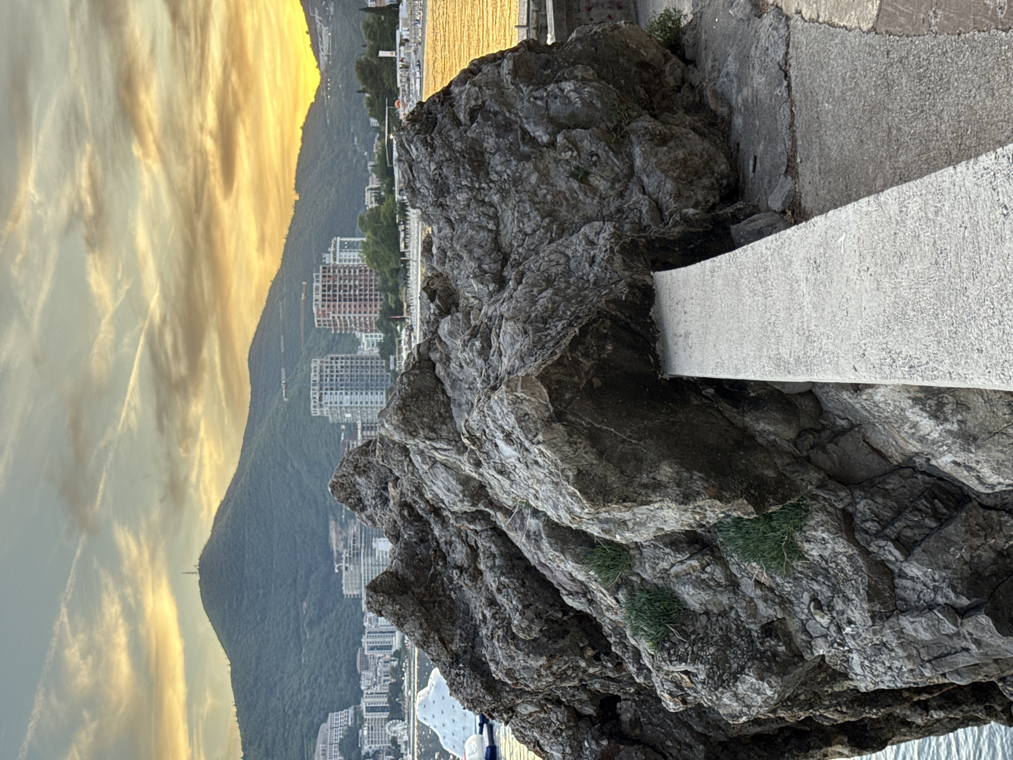 The image depicts a large, weathered rock formation in the foreground, with a narrow concrete walkway extending from its base. Behind the rock, the city of Budva, Montenegro, is visible, characterized by numerous buildings and a hill covered in greenery. The sunset casts a warm golden glow across the sky, illuminating wispy clouds and the cityscape. The sea is a calm, reflective surface reflecting the sunset colors. The overall scene is peaceful and picturesque, capturing the beauty of the Budva coastline at dusk.
