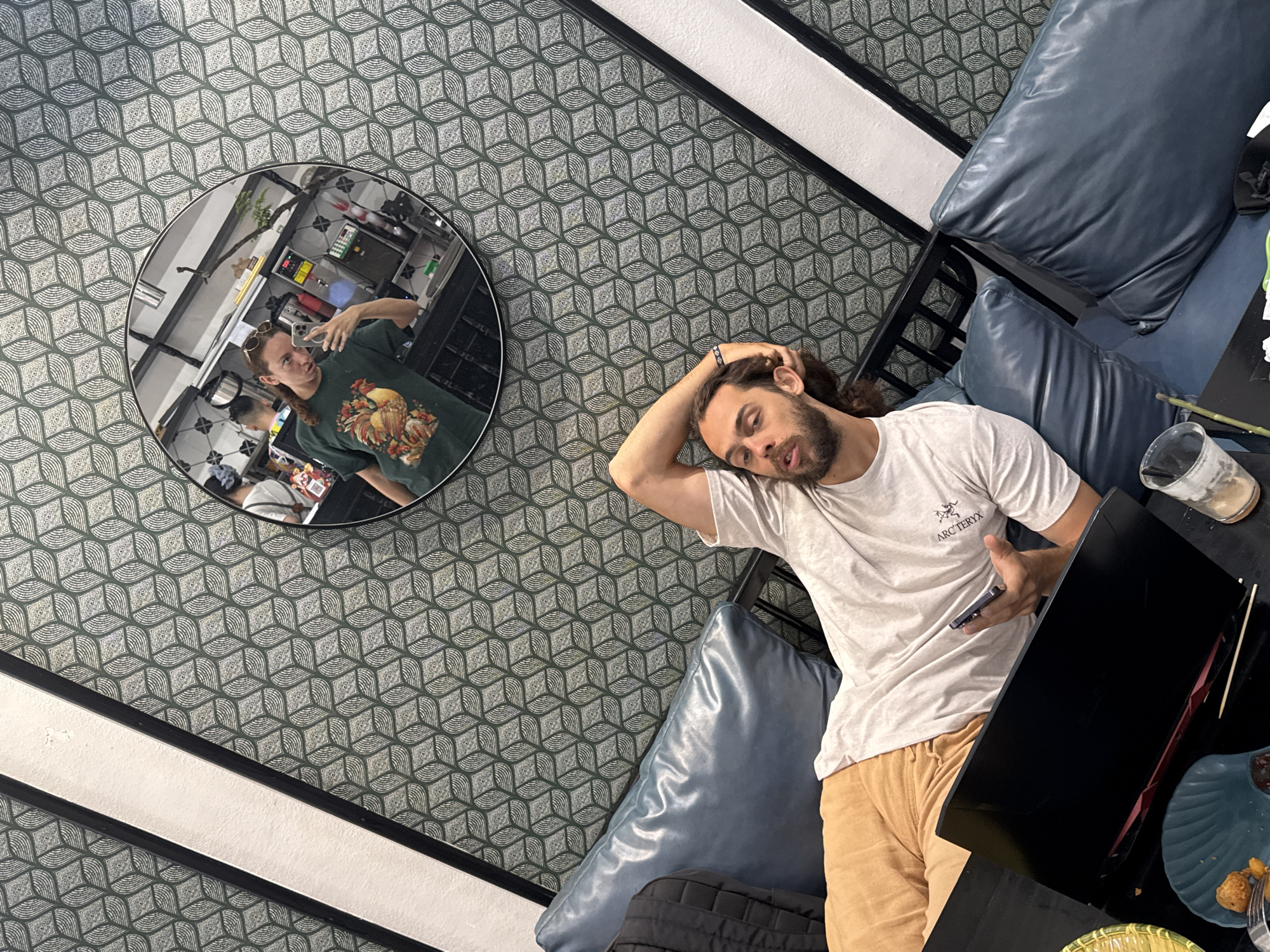 The image captures an indoor scene, likely a cafe or lounge in Phố Ràng, Vietnam, taken from an elevated and tilted perspective. The dominant feature of the room is a patterned wallpaper with a repeating geometric design of green and white abstract shapes.
In the foreground, a man with long, dark hair and a beard is reclining on a dark blue faux leather sofa. He is wearing a white t-shirt with "ARC'TERYX" visible on the chest, and light tan shorts. His right hand is behind his head, and he is holding a mobile phone in his left hand, appearing relaxed or contemplative.
To the left of the man, a circular mirror reflects another person taking a selfie. This individual, wearing sunglasses on their head and a dark green t-shirt with a large, colorful rooster graphic, holds a phone up to capture their reflection. The mirror also reveals parts of the interior behind them, possibly a service or kitchen area with metal shelving and various containers.
On a black table beside the man, there is a clear glass with a straw, a blue plate containing some fried food items and a fork, and part of a yellow-green woven basket with more food. The overall ambiance suggests a casual, relaxed gathering during the daytime.