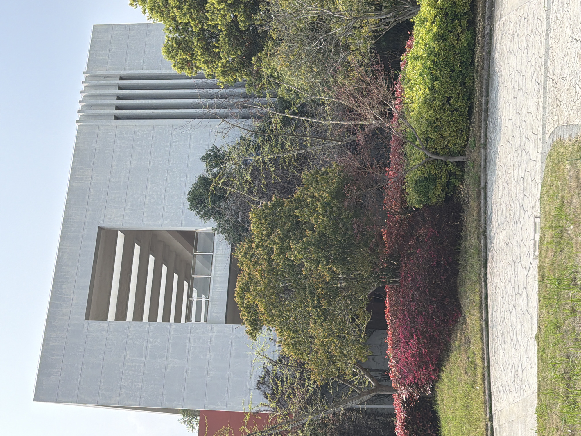 A modern building with a unique architectural design is captured from a ground-level perspective. The building, constructed from concrete, stands prominently on the left side of the frame. Its features include horizontal concrete slats at the top and a large, open rectangular section with vertical slats at the bottom.
The right side of the image is dominated by a combination of trees, bushes, and greenery. A paved path borders the greenery, suggesting a walkway or pathway. The scene appears to be set in an urban or suburban environment, with a clear sky in the background.
There is no indication of Jiangning District, China, nor is there any detail on people, subjects, objects, actions, or interactions.