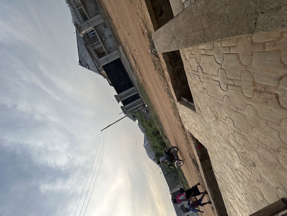 An outdoor scene shows buildings under construction and a dirt road. One building displays a patterned concrete wall with a window opening. A motorcycle is parked on the side of the road, with several children standing nearby. Overhead power lines stretch across a cloudy sky. The perspective is slightly skewed, with a tilted horizon.