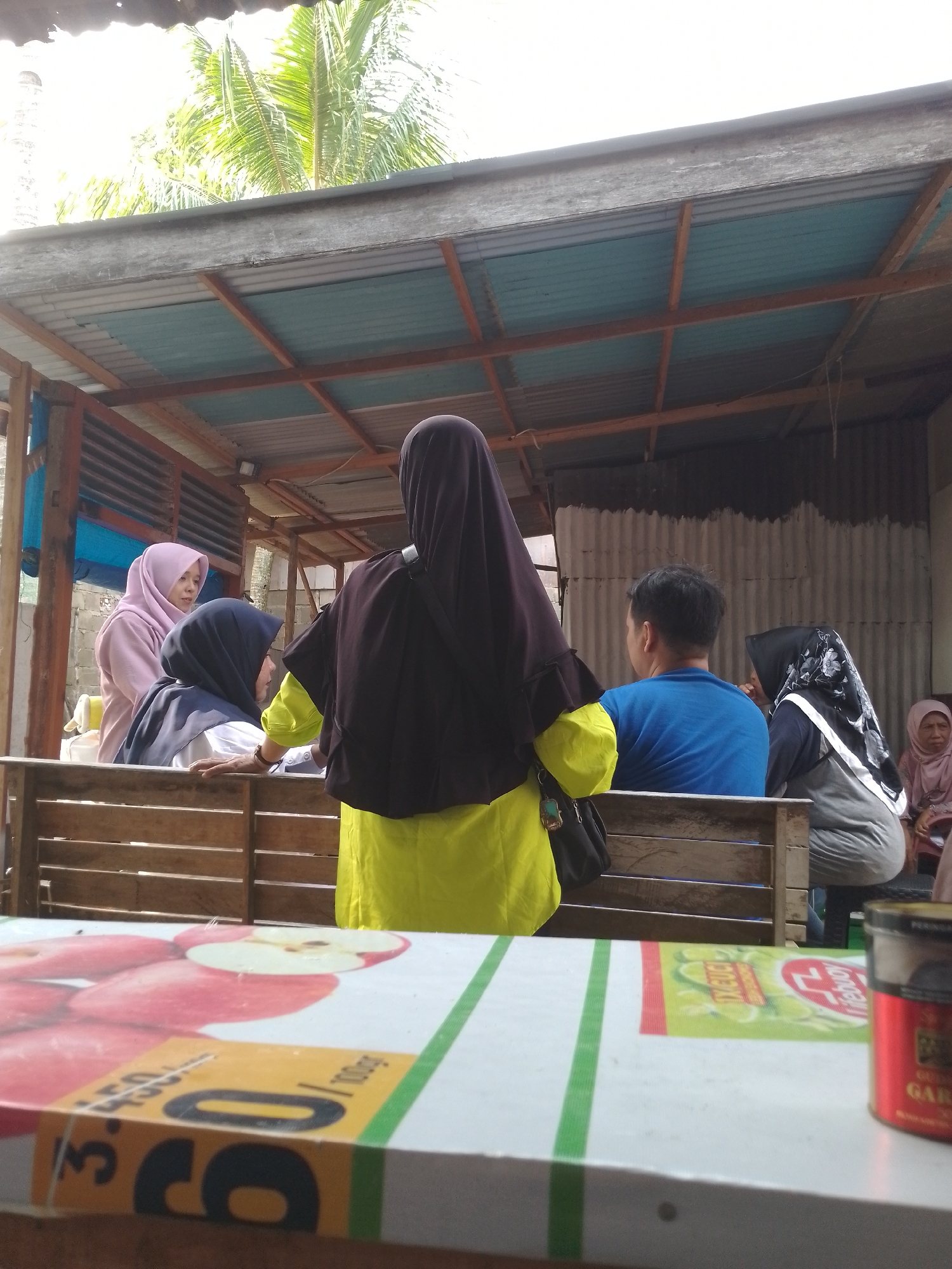 The image depicts an outdoor scene under a simple shelter, possibly a food stall or a resting area, in the city of Padang, Indonesia. In the foreground, a table is visible, adorned with an image of apples and some packaging, including a red tin with Indonesian writing. Behind the table, several people are present.
Two women wearing hijabs are positioned on the left side, one in a pink hijab and the other in a dark blue hijab. A third person, facing away from the camera and wearing a dark hijab and a bright yellow garment, stands in the center. To the right, a man in a blue t-shirt is seated, and further to the right, another person in a black and white patterned hijab is also seated. An elderly woman in a pink hijab is partially visible on the far right.
The shelter features a corrugated metal roof supported by wooden beams. The background shows a corrugated metal wall and some greenery, including palm fronds, suggesting a tropical environment. The overall atmosphere appears casual and possibly social, with people gathered in a relaxed setting.