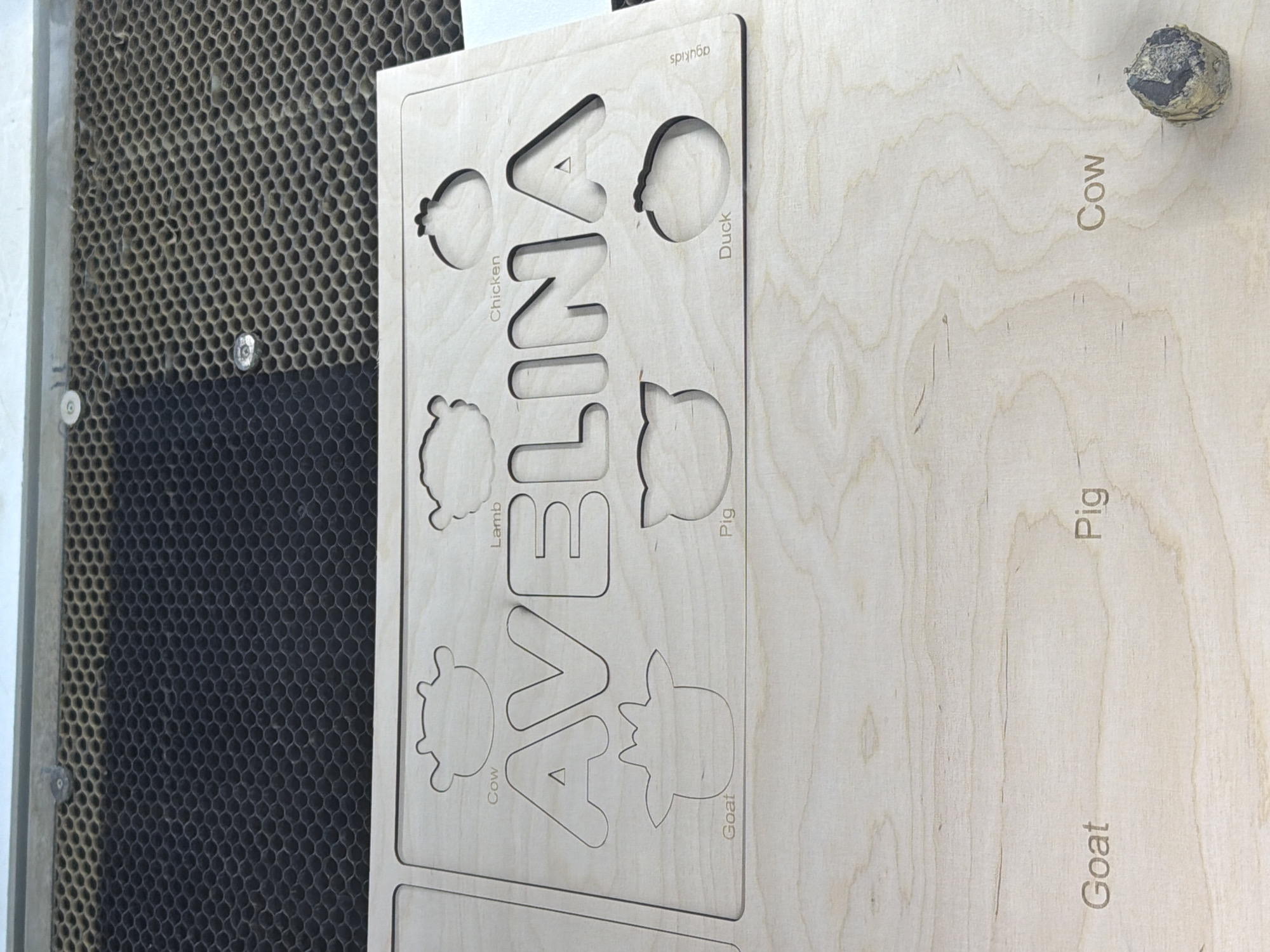 A wooden puzzle board with the name "AVELINA" cut out in large letters is shown. Around the name, there are outlines of farm animals, each with its name labeled below. The animals include a cow, lamb, chicken, goat, pig, and duck. The puzzle appears to be a learning toy for children. The background shows a honeycomb-like surface, likely a laser cutter bed, suggesting the puzzle was recently made. A small, dark object, possibly a piece of wood debris, is visible in the bottom right corner. The scene is indoors and well-lit, with the focus on the puzzle itself.