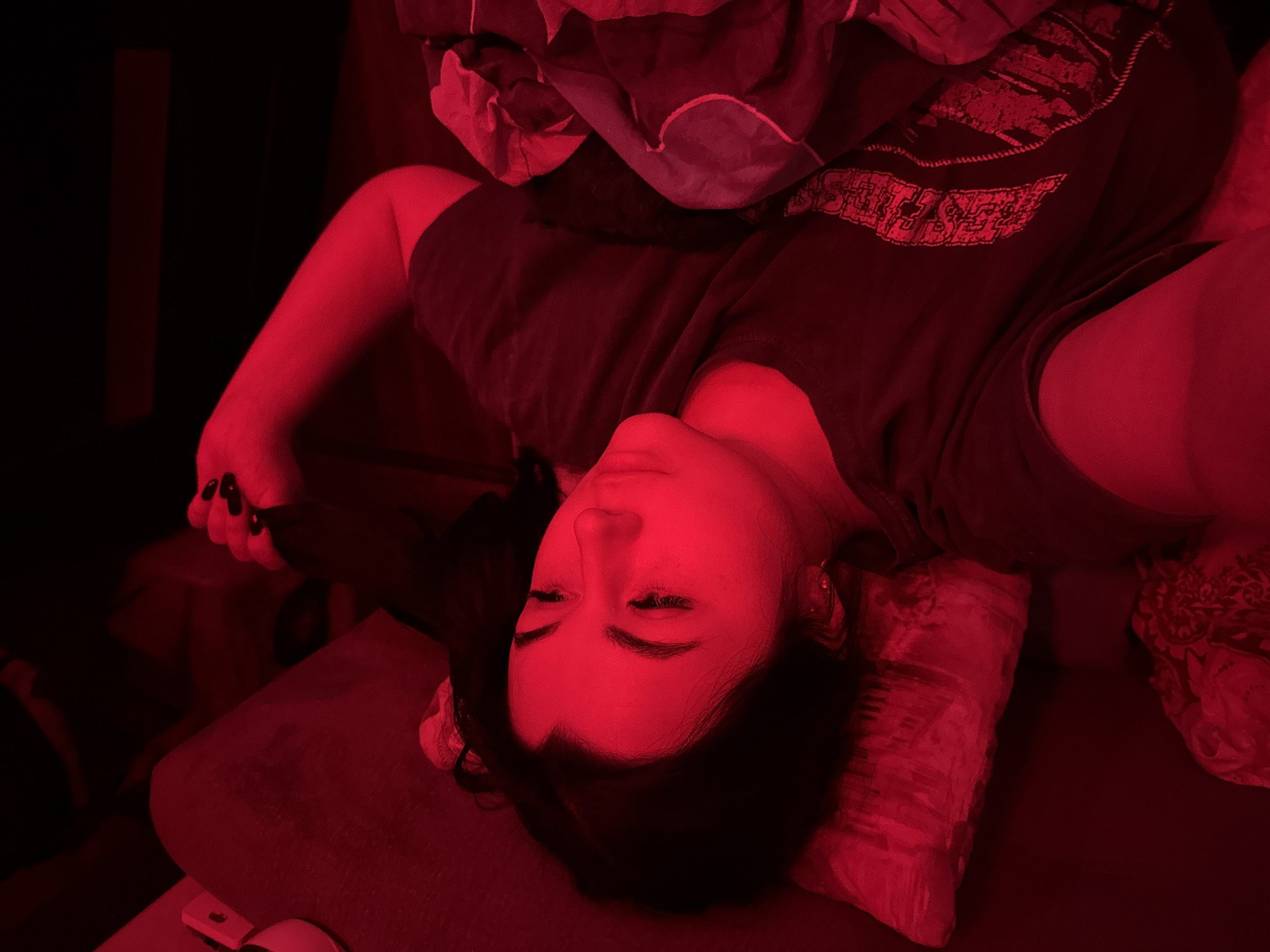 A young woman with long dark hair is lying on a pillow in what appears to be a bed. She is wearing a dark-colored t-shirt with a graphic design on the front. The lighting is predominantly red. She is holding a black item in her right hand. The location is not specified in the image. The city of Chaikovsky, Russia is also not in the image.