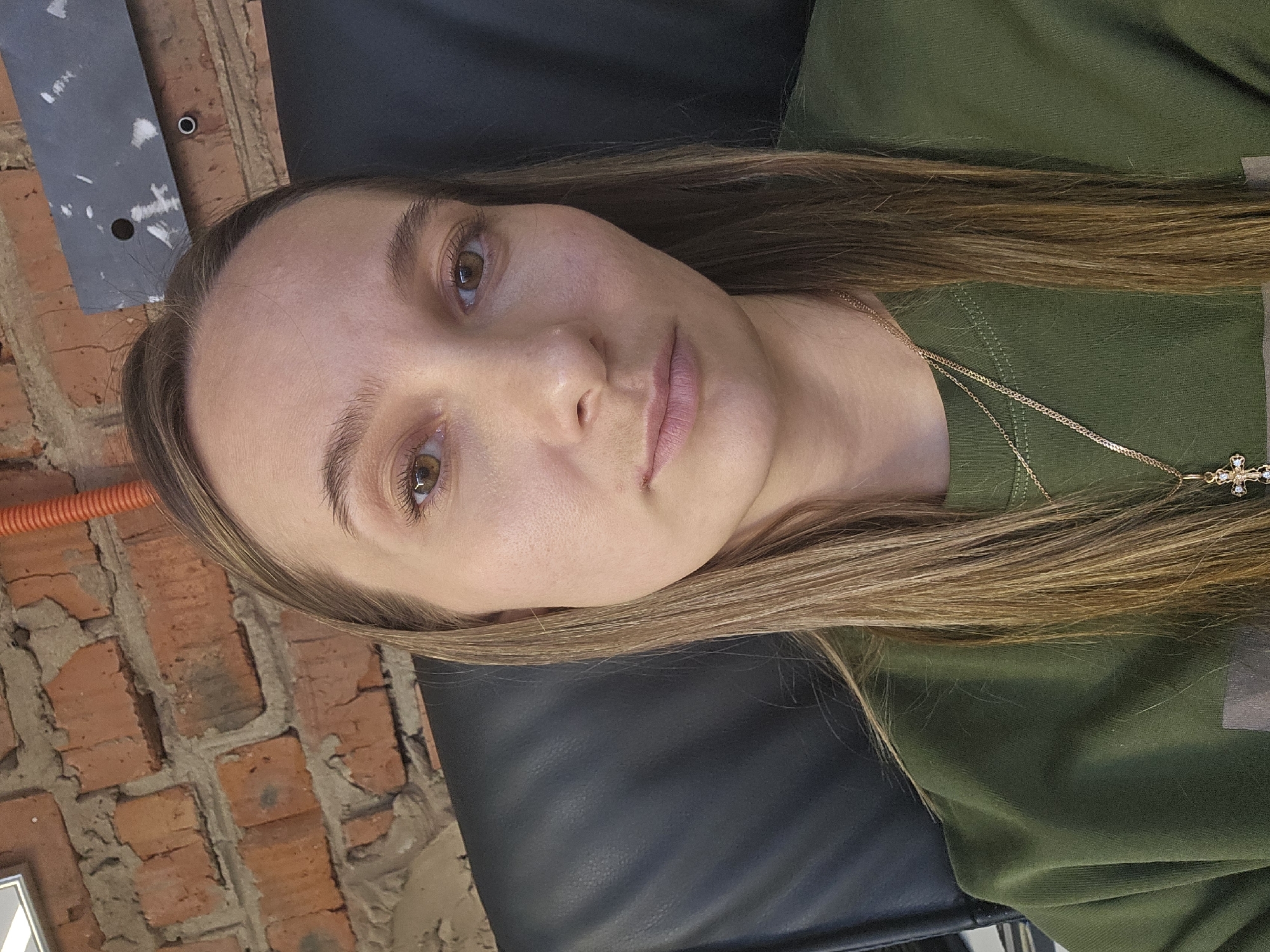 A woman with light brown hair and eyes is lying on a black leather surface. She is wearing a green shirt and a gold necklace with a cross pendant. The background includes a brick wall with some electrical conduit visible. The image does not provide enough information to confirm that the location is Chaikovsky, Russia.