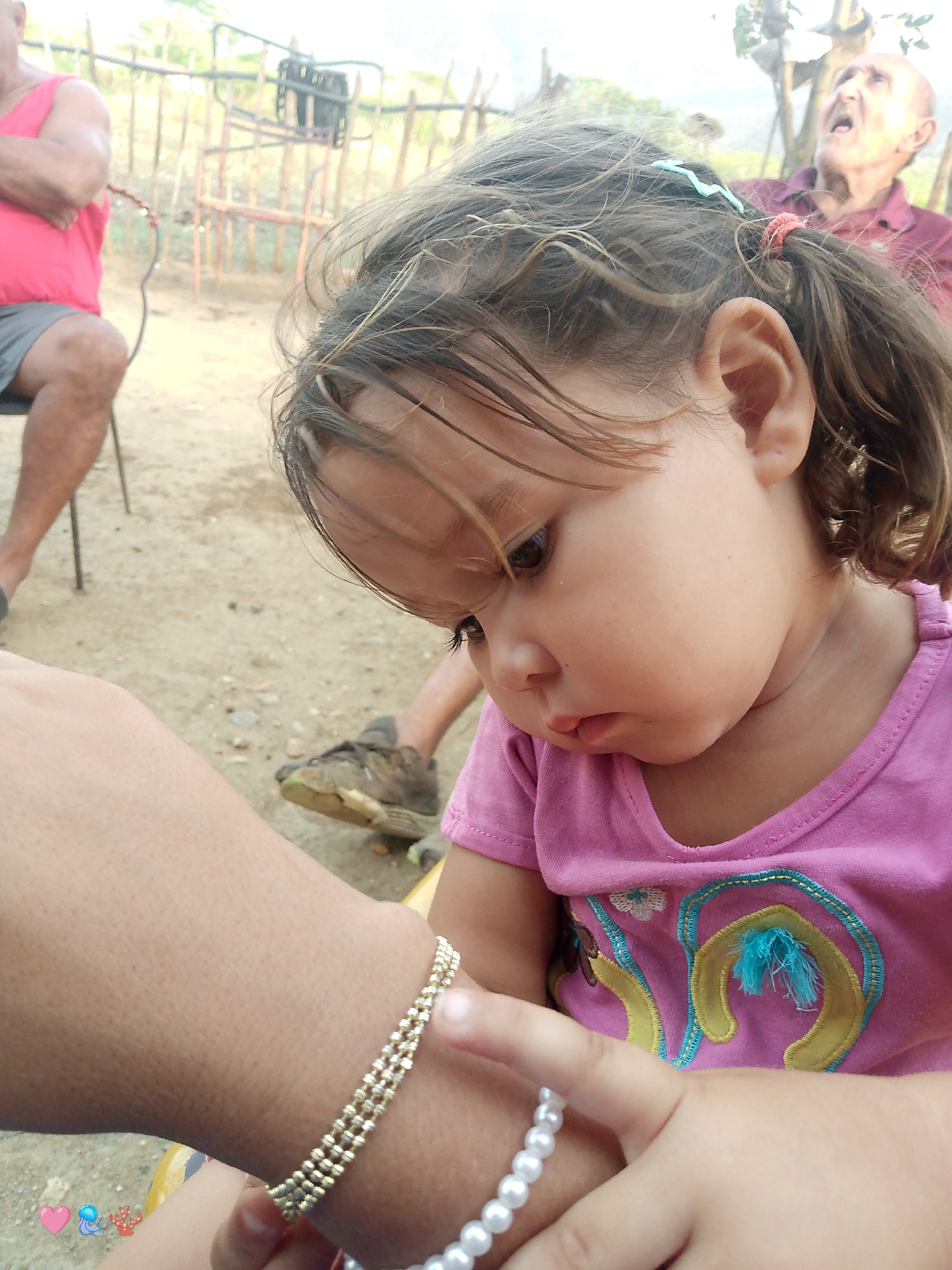 In El Cercado, Venezuela, a close-up shot focuses on a young child wearing a pink shirt with embroidered details. The child's head is tilted downwards, revealing a portion of their face and hair, which is tied back with a red elastic band and a blue clip. The child's left arm, adorned with a gold beaded bracelet, is prominent in the foreground. A second, pearl bracelet is visible on the child's right hand. In the background, an older person in a red tank top and grey shorts is seated, and another elderly person in a purple shirt is visible in the upper right corner of the image, appearing to be speaking. The setting appears to be outdoors, with a dirt ground and a wooden fence in the background, suggesting a rural or semi-rural environment.