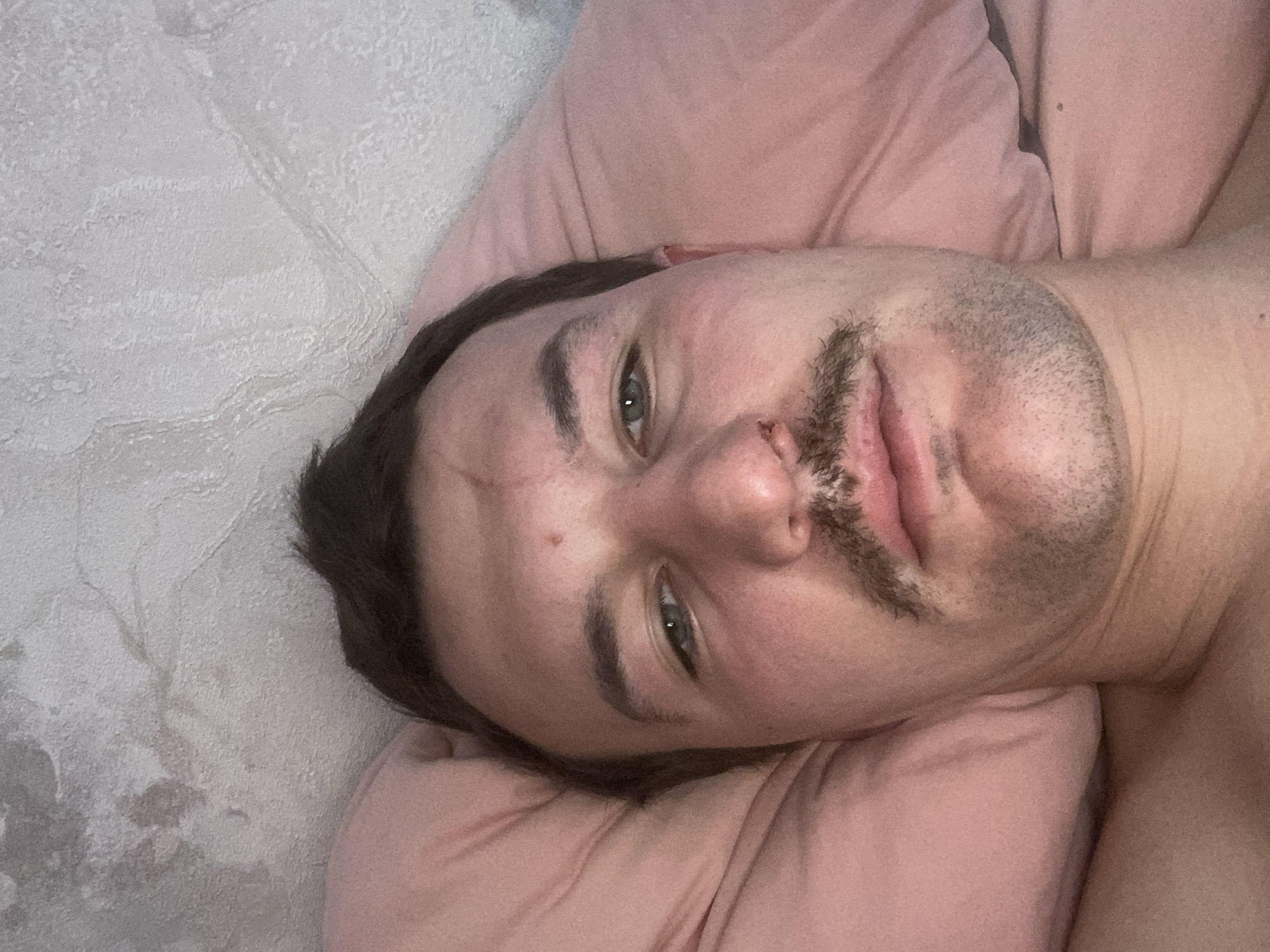 A man is lying on a pink pillow. His head is resting on the pillow, and his body is angled to the right. He has short dark hair, a mustache, and stubble. His eyes are open and he appears to be looking at the camera. The background consists of a grey wall. The city of Sochi, Russia is not discernable from the image.