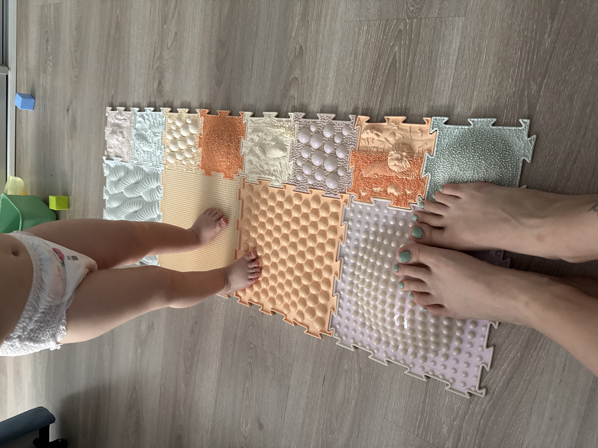 The image shows a top-down view of two individuals standing on a colorful textured puzzle mat placed on a wooden floor. The individual on the left appears to be a baby or toddler, wearing a diaper. The individual on the right is an adult, with their feet visible. The puzzle mat consists of various textured squares. In the background, there are scattered toys and furniture. The scene takes place indoors, likely in a private residence. There is no information to determine the location as Moscow, Russia.