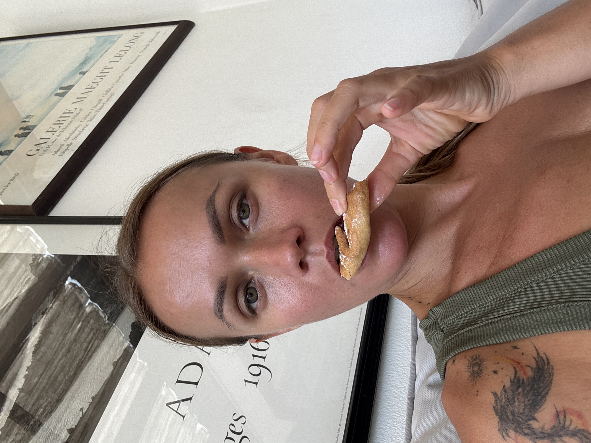 A woman is captured in a close-up selfie as she eats a pastry. The woman has blonde hair pulled back and green eyes. She is wearing a green tank top. In the background, there are two framed pieces of art hanging on a white wall. To the left, a large abstract painting with black and white brush strokes. To the right, a framed poster with text and a silhouette of people walking on a beach. The scene appears to be indoors. There is no visible indication of the location being Baan Mae Nam, Thailand.