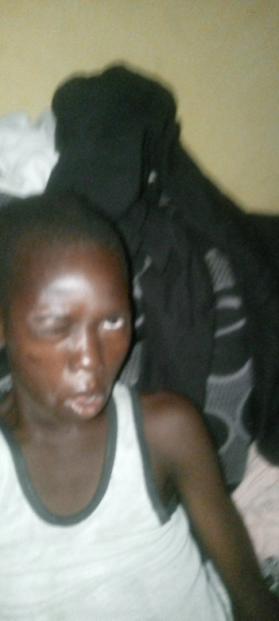 A blurry, low-resolution image captures a person, likely a child or young adult, from the chest up. The individual has dark skin and is wearing a white tank top with dark trim around the neck and armholes. Their face is somewhat obscured by motion blur and poor lighting. Their short, dark hair is visible at the top of their head. The eyes appear partially closed or squinting; one eye shows a bright white reflection or distinct light coloring. The mouth is slightly open. The background consists of a light-colored, possibly beige or pale yellow, wall on the left and a dark, indistinct mass of fabric or clothing on the right, suggesting an indoor setting. The lighting appears to be low or artificial, causing bright reflections on the person's forehead and nose. No text is visible.