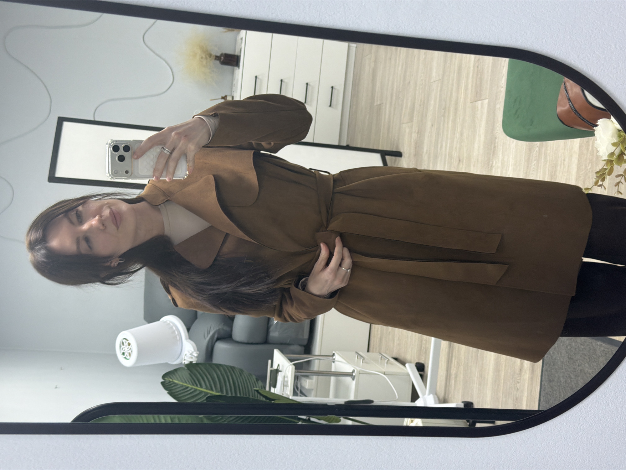 The image shows a woman taking a selfie in a mirror. She is wearing a brown trench coat. The reflection shows a room with a sofa, a dresser, a plant, and a chair. The location of the image is not identifiable as Perm, Russia.